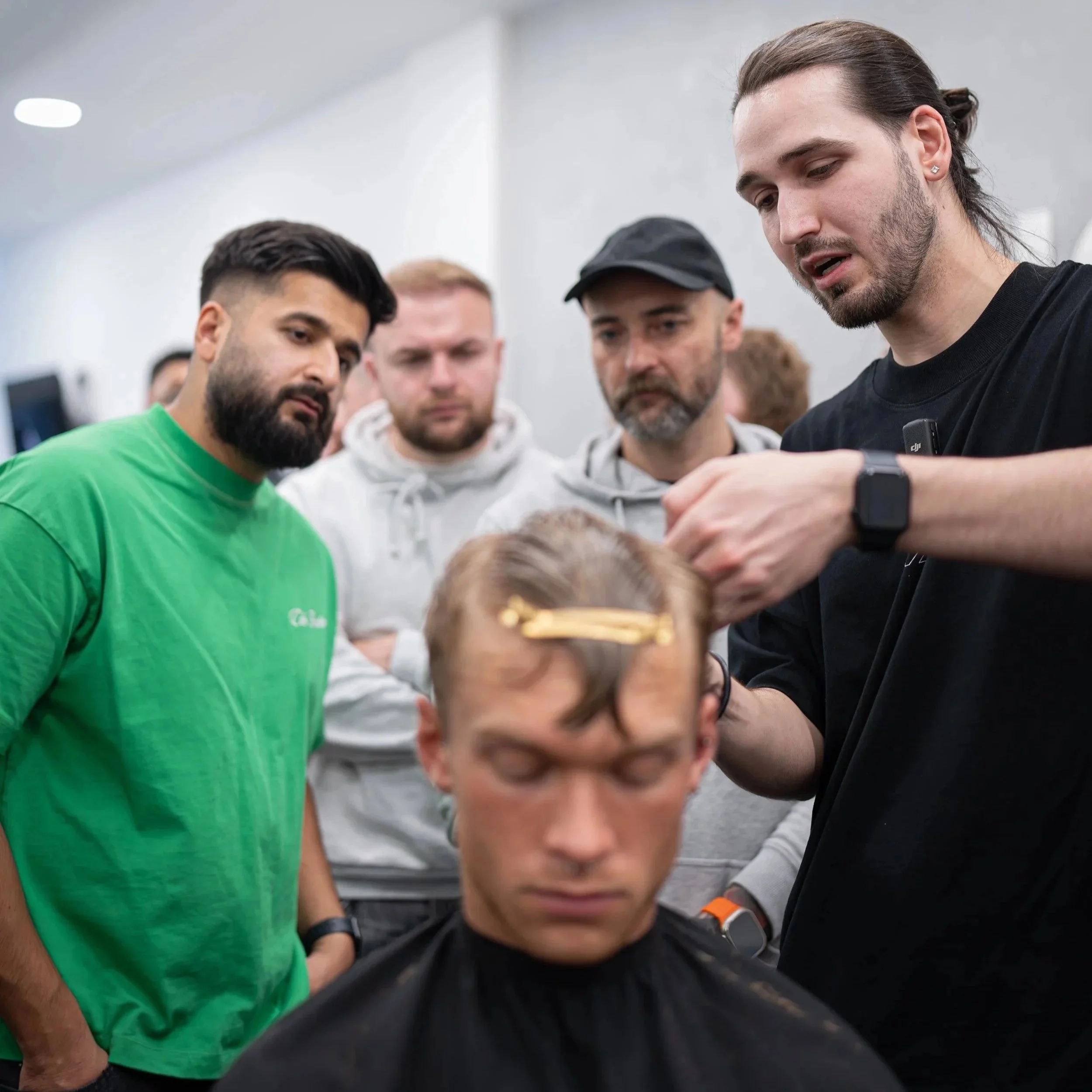 A barber in black shirt giving a haircut to a man with glasses in a salon, while four onlookers watch closely.