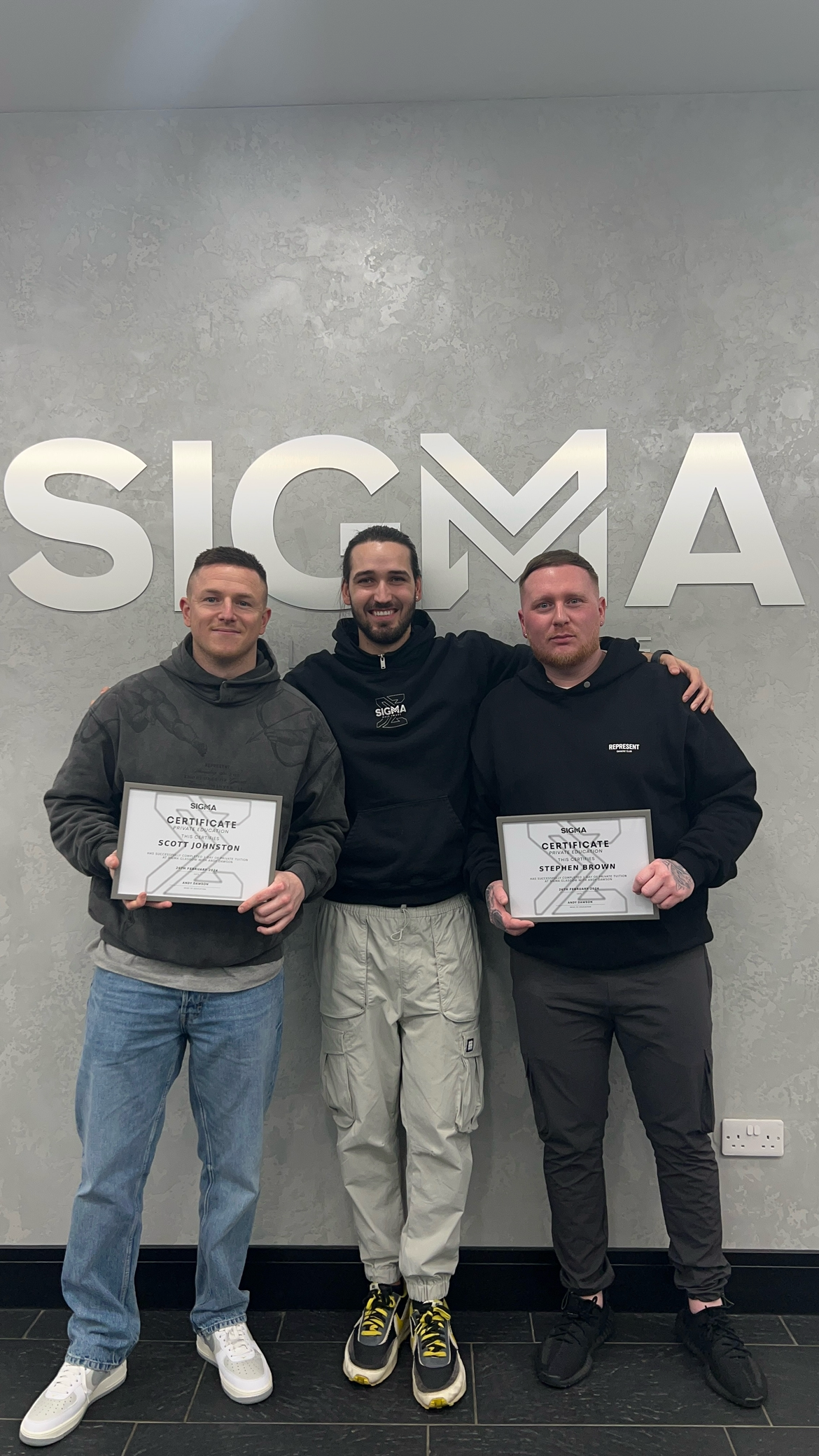Three men standing in front of a wall with a large SIGMA sign, two of them holding certificates, and smiling at the camera.