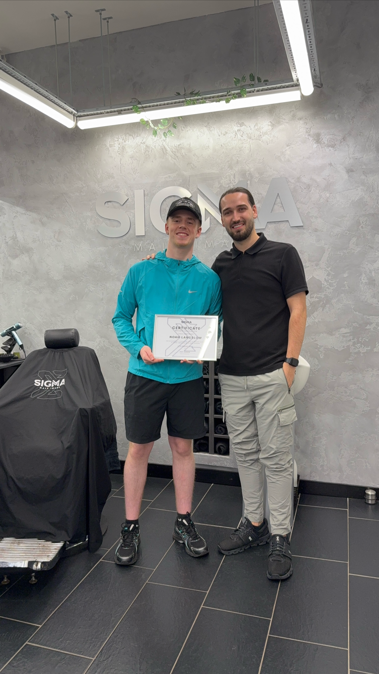 Two men standing in a gym, one holding a diploma or certificate, smiling. The man on the left wears a blue jacket and black shorts, the other man on the right wears a black polo shirt and light gray pants. The wall behind them has the logo 'SIGMA'.