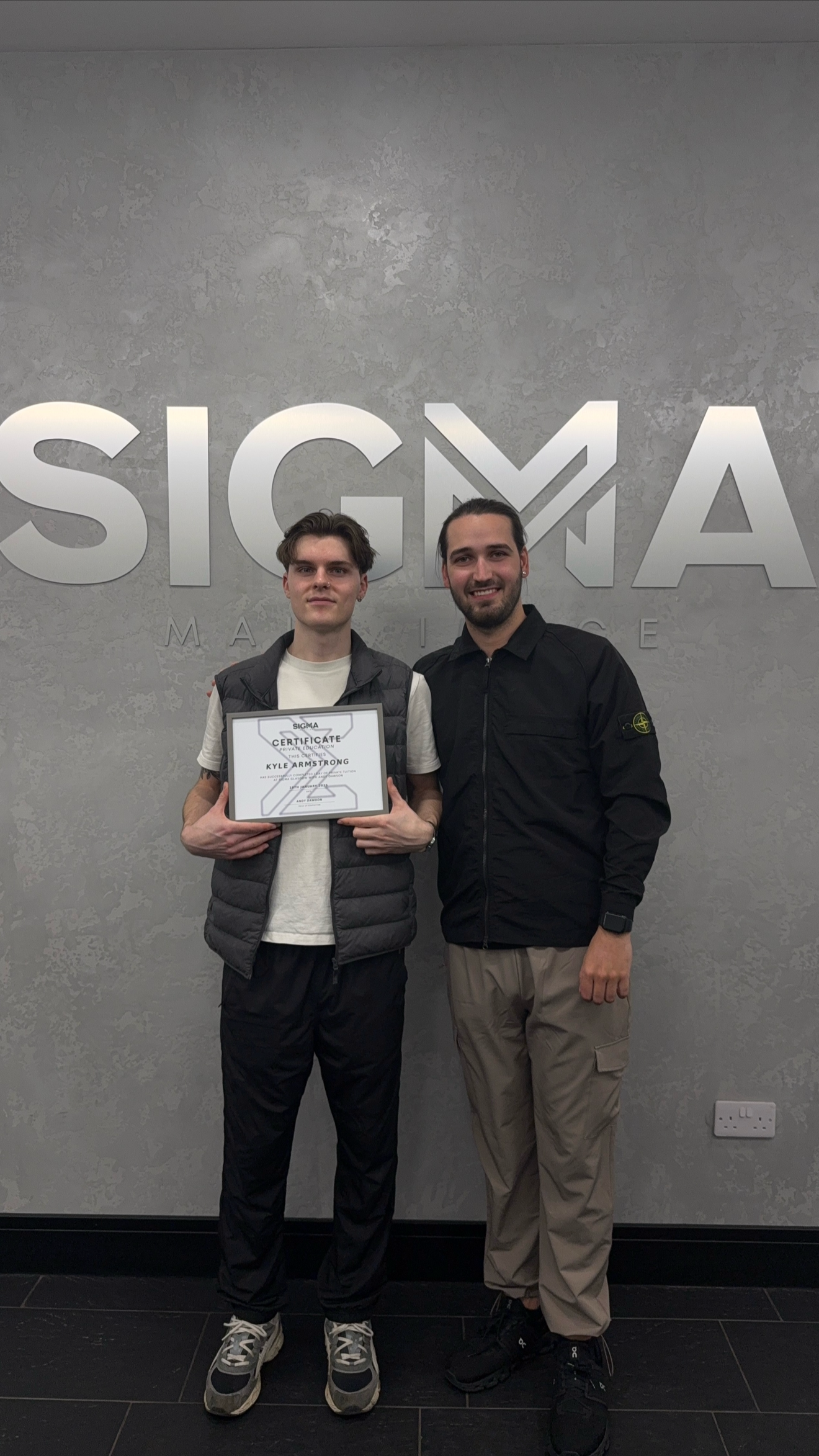 Two young men standing in front of a gray wall with the Sigma Marine logo. One is holding a framed certificate with the name Kyle Armstrong. Both are smiling, and the man on the right is wearing a black jacket and beige pants.