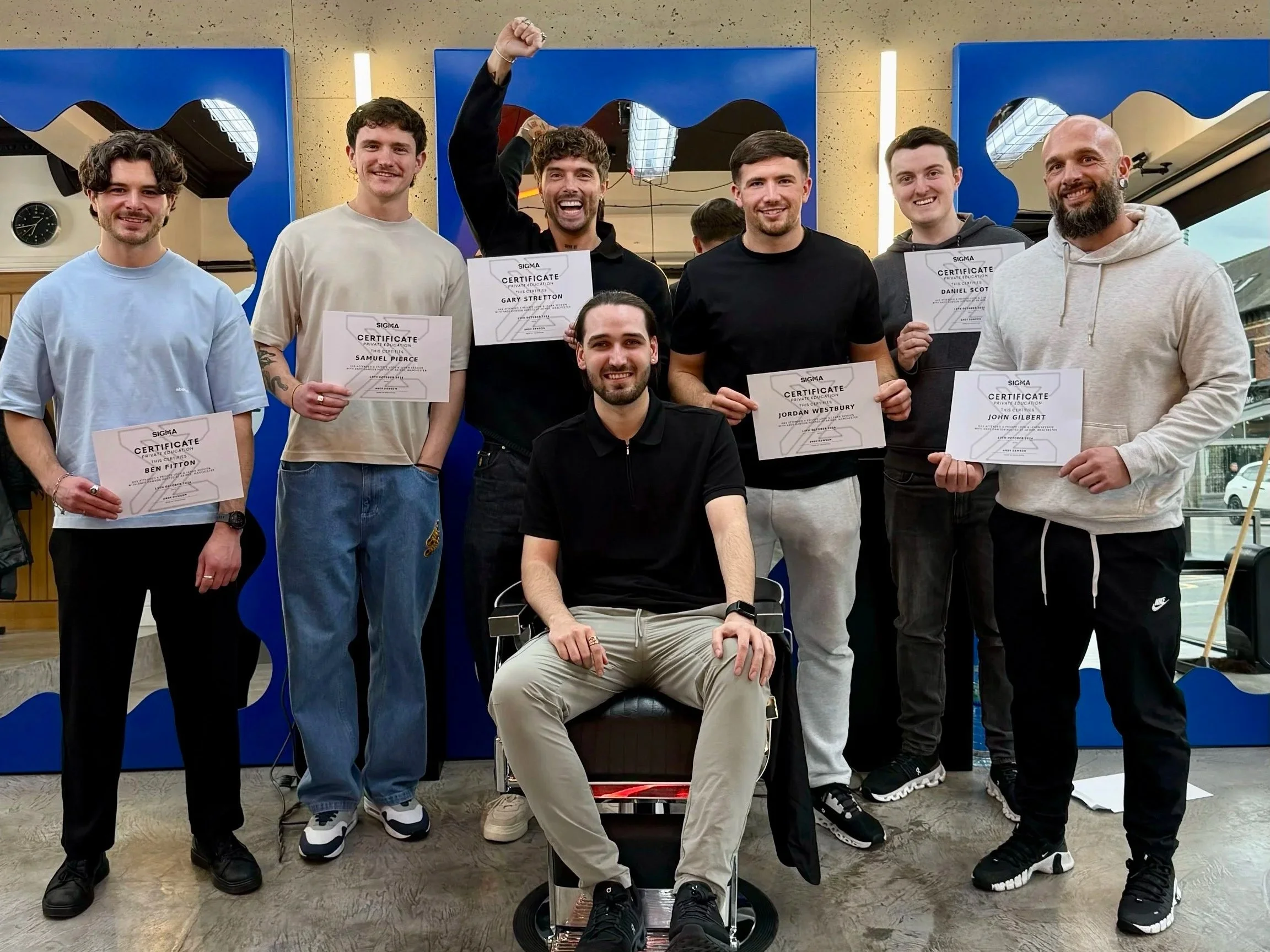 Group of seven young men celebrating an achievement, holding certificates, posing indoors with a man seated in front of them, blue decorative panels behind, one man raising a fist in victory, smiling.