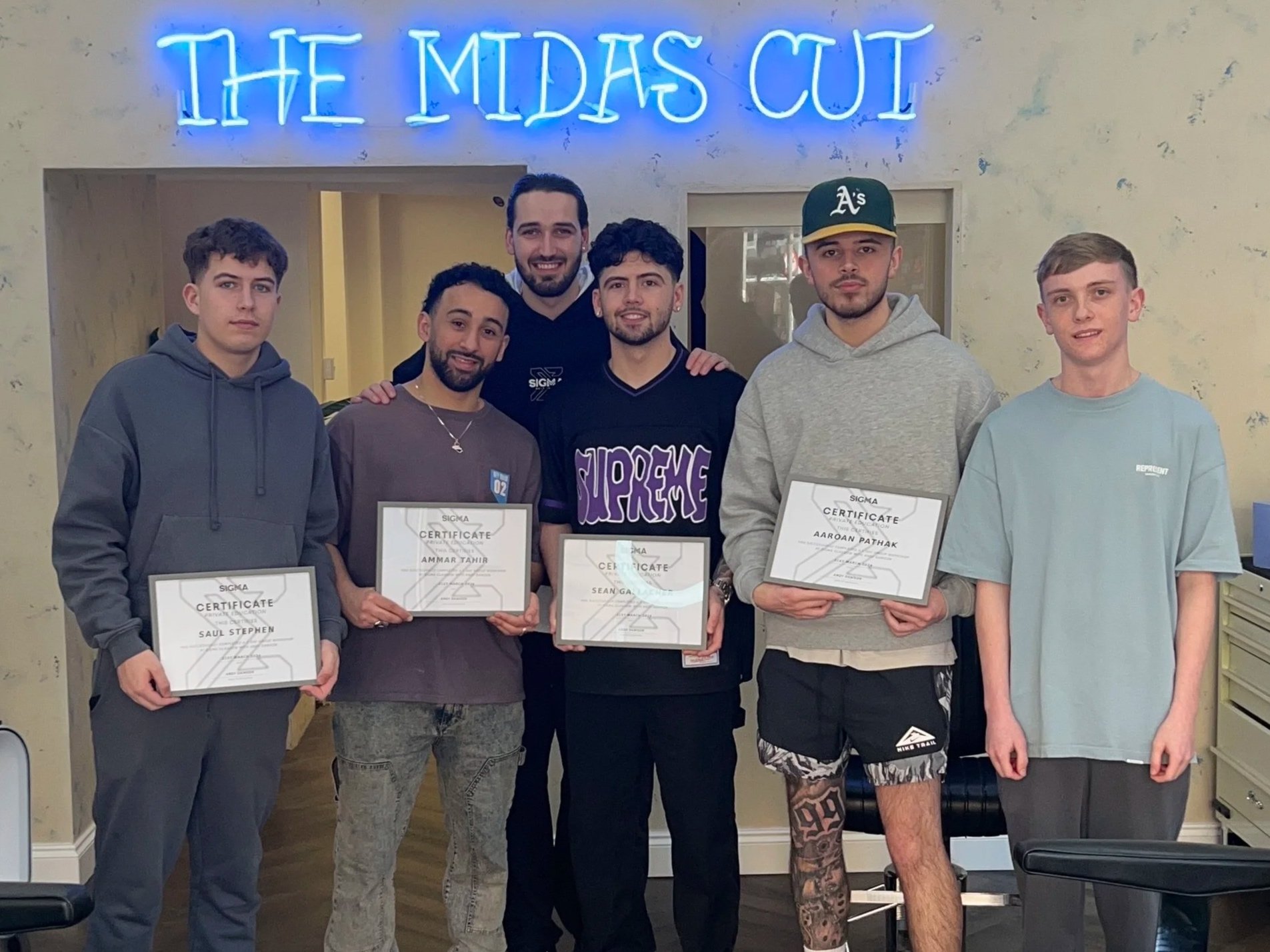 Six young men standing in a room with a beige wall and a bright blue neon sign that reads 'THE MIDAS CUT'. Four of them are holding certificates. The group appears to be celebrating or at an award ceremony.