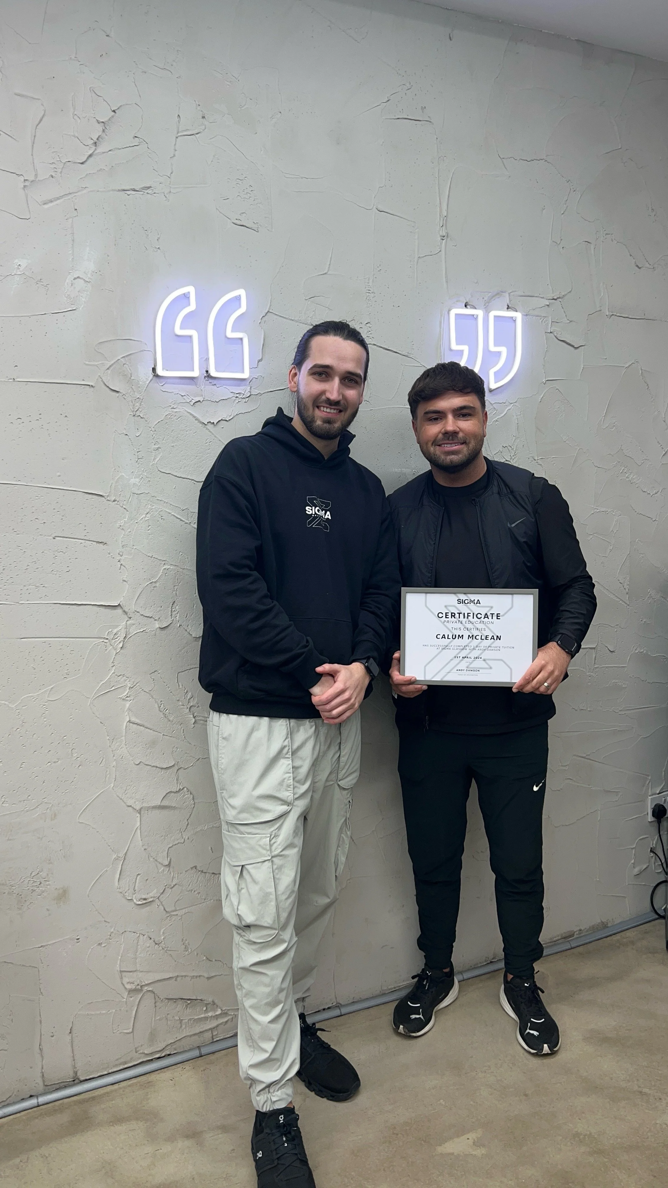 Two men standing indoors against a textured wall with illuminated quote marks. The man on the right is holding a framed certificate, and both are smiling.