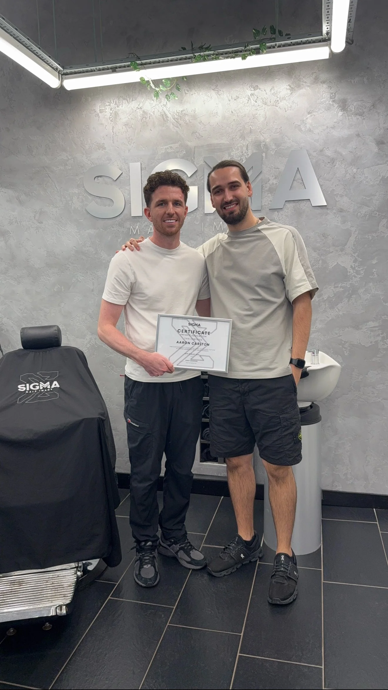 Two men in casual clothing standing inside a modern room with a gray textured wall, holding a certificate, smiling at the camera, with Sigma logo and a barber chair covered with a Sigma cloth nearby.