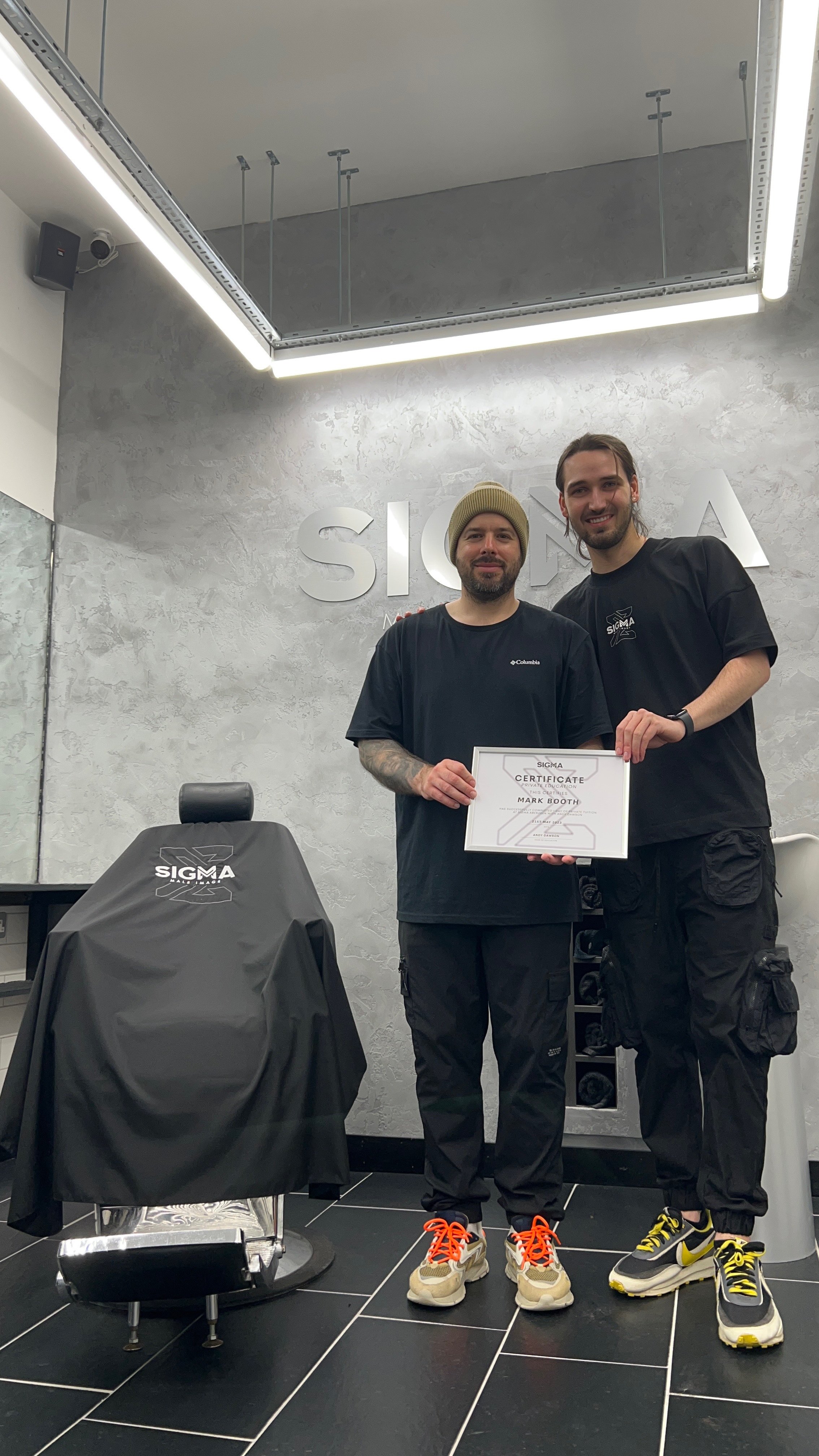 Two men standing in a modern salon, one holding a certificate, both smiling, with salon equipment and Sigma branding in the background.