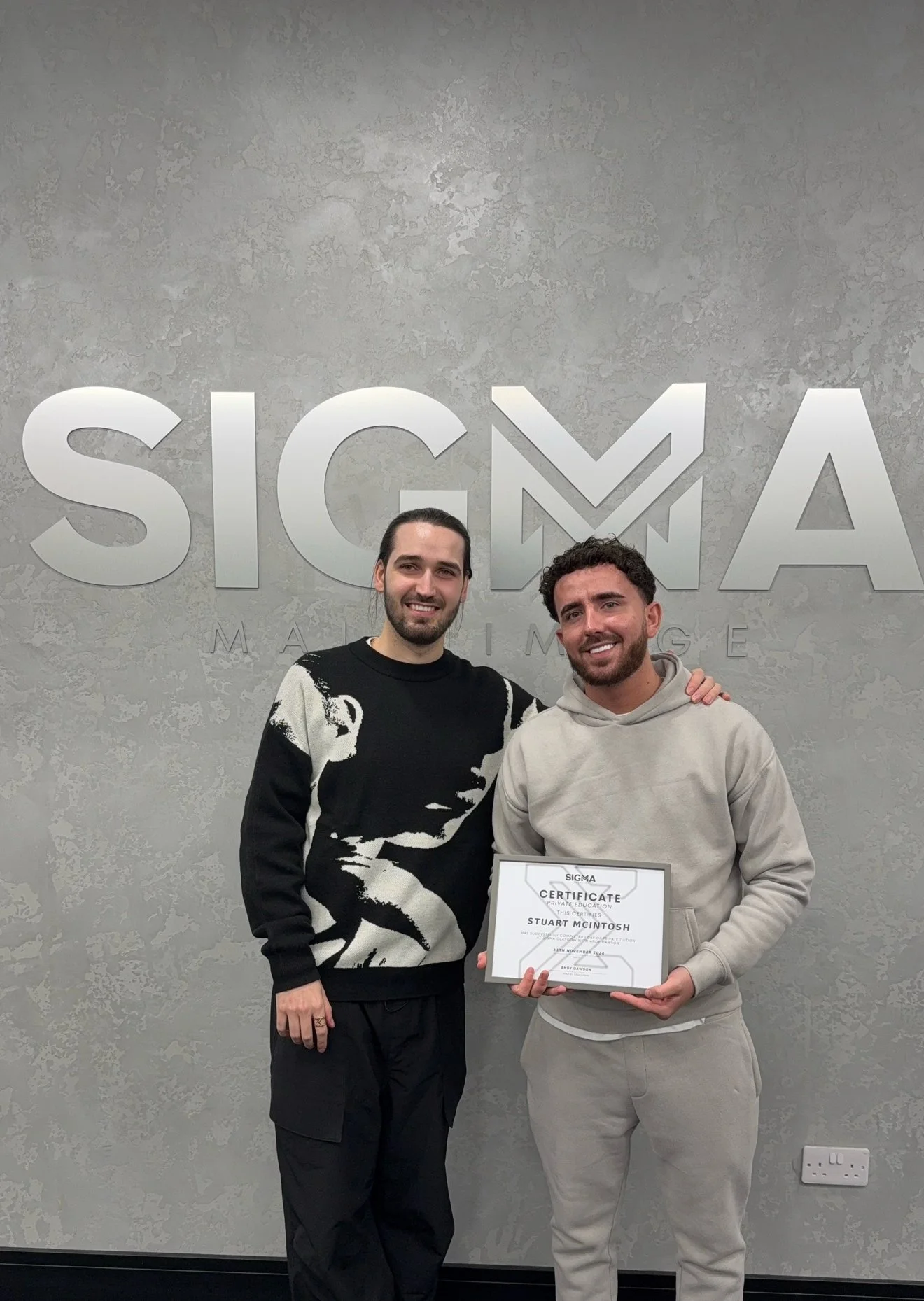 Two men stand in front of a wall with the SIGMA logo; one holds a certificate of achievement, and both are smiling.