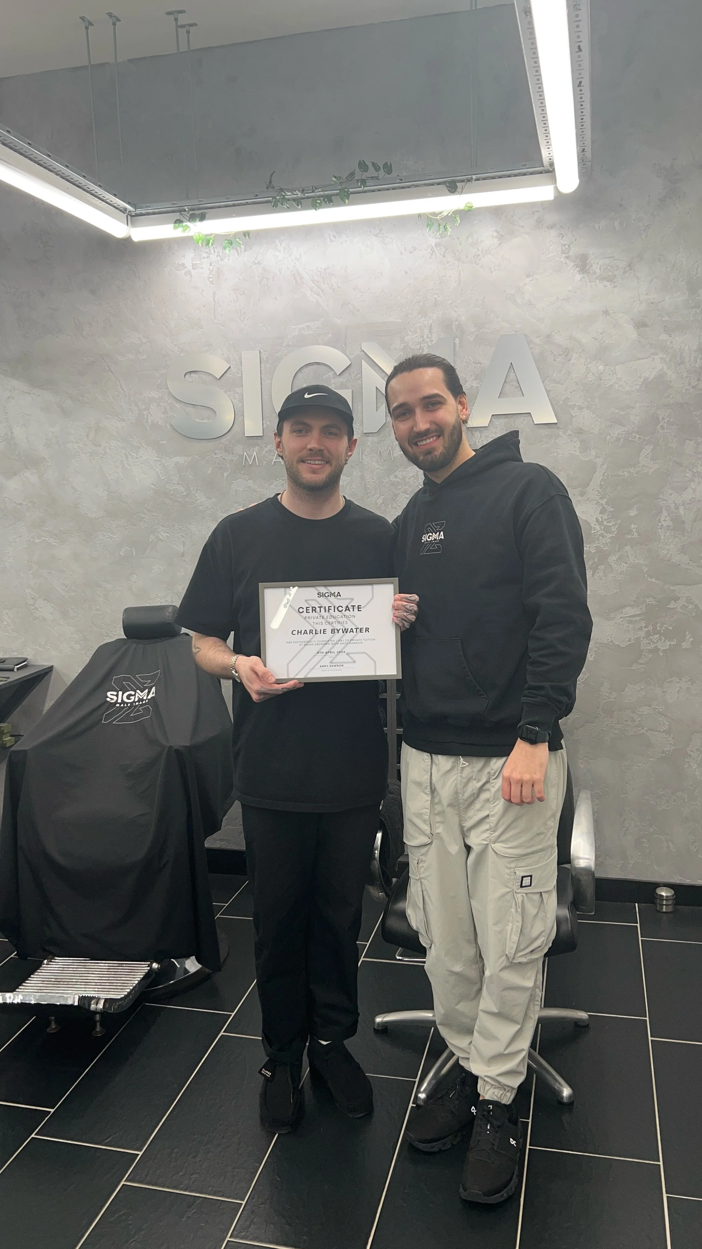 Two men standing in a room with grey textured walls, one holding a certificate. The room has a Sigma logo on the wall behind them and a covered chair to the side. Both men are smiling, dressed casually in black and grey.