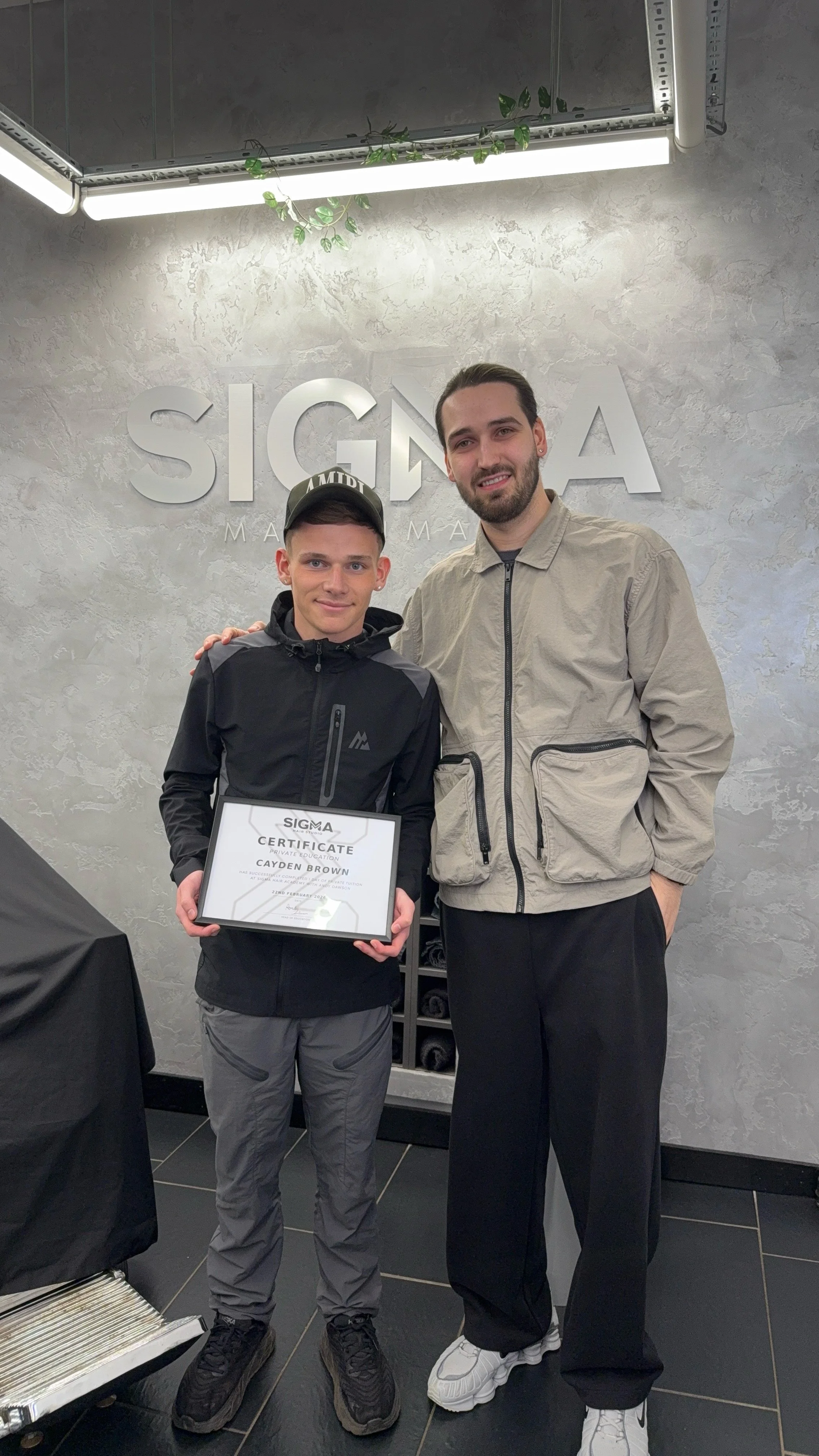 Two men standing together inside a building, one holding a certificate. The background features a wall with the words "SIGMA" and some greenery hanging from the ceiling.