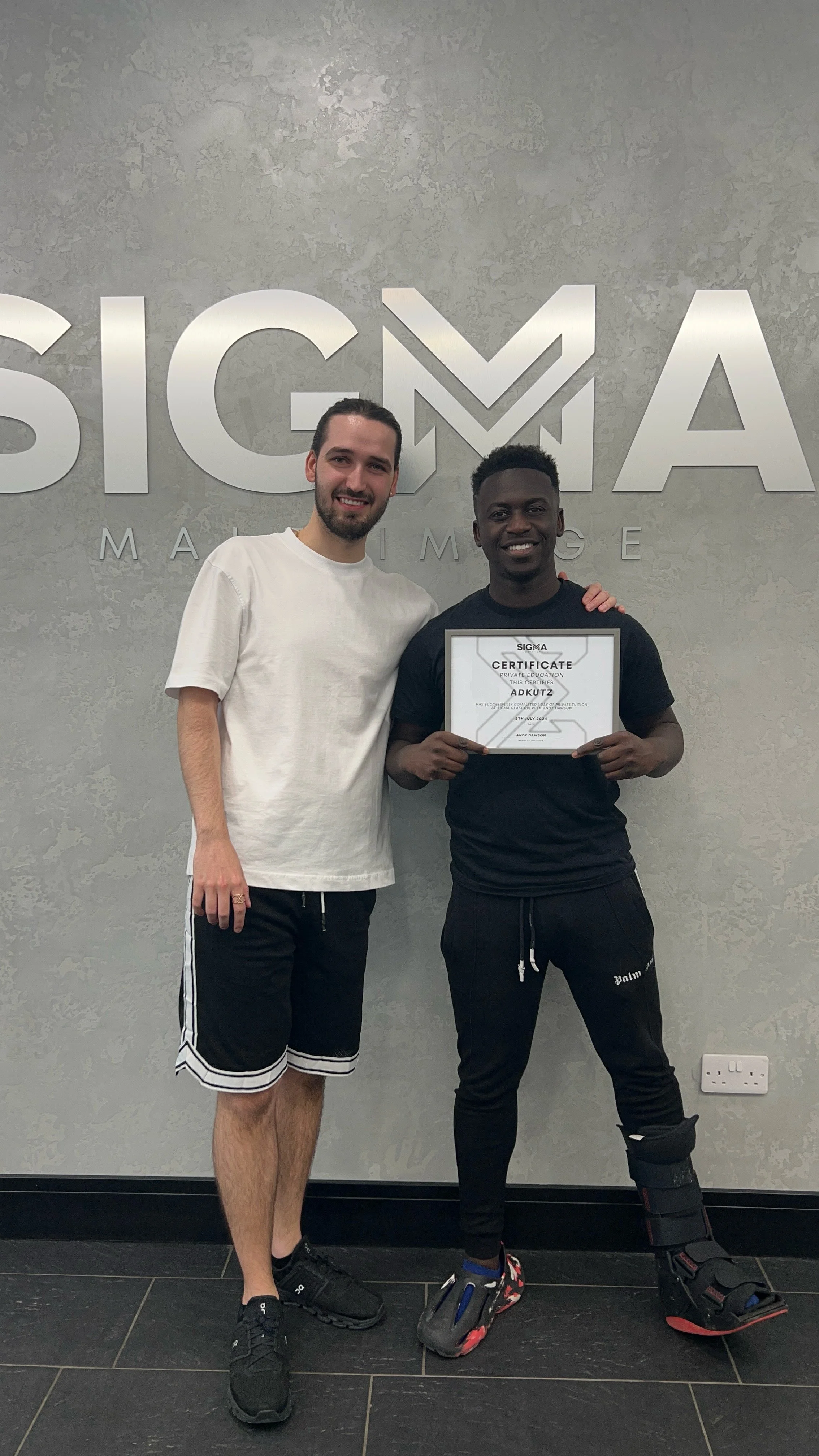 Two men standing in front of a wall with the SIGMA logo, smiling, one holding a certificate, the other with his arm around him.