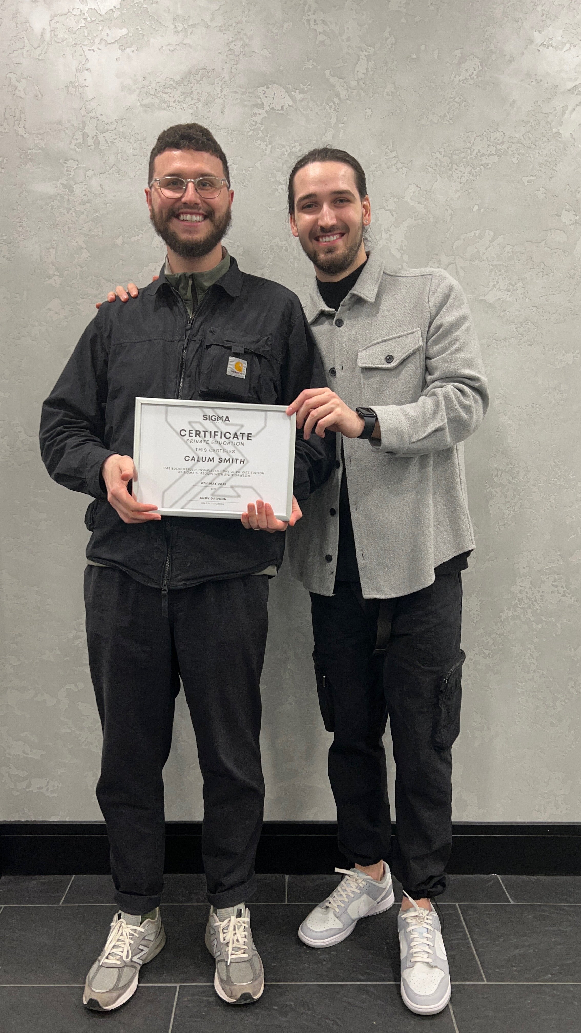 Two young men smiling; one holding a certificate, standing against a gray textured wall.