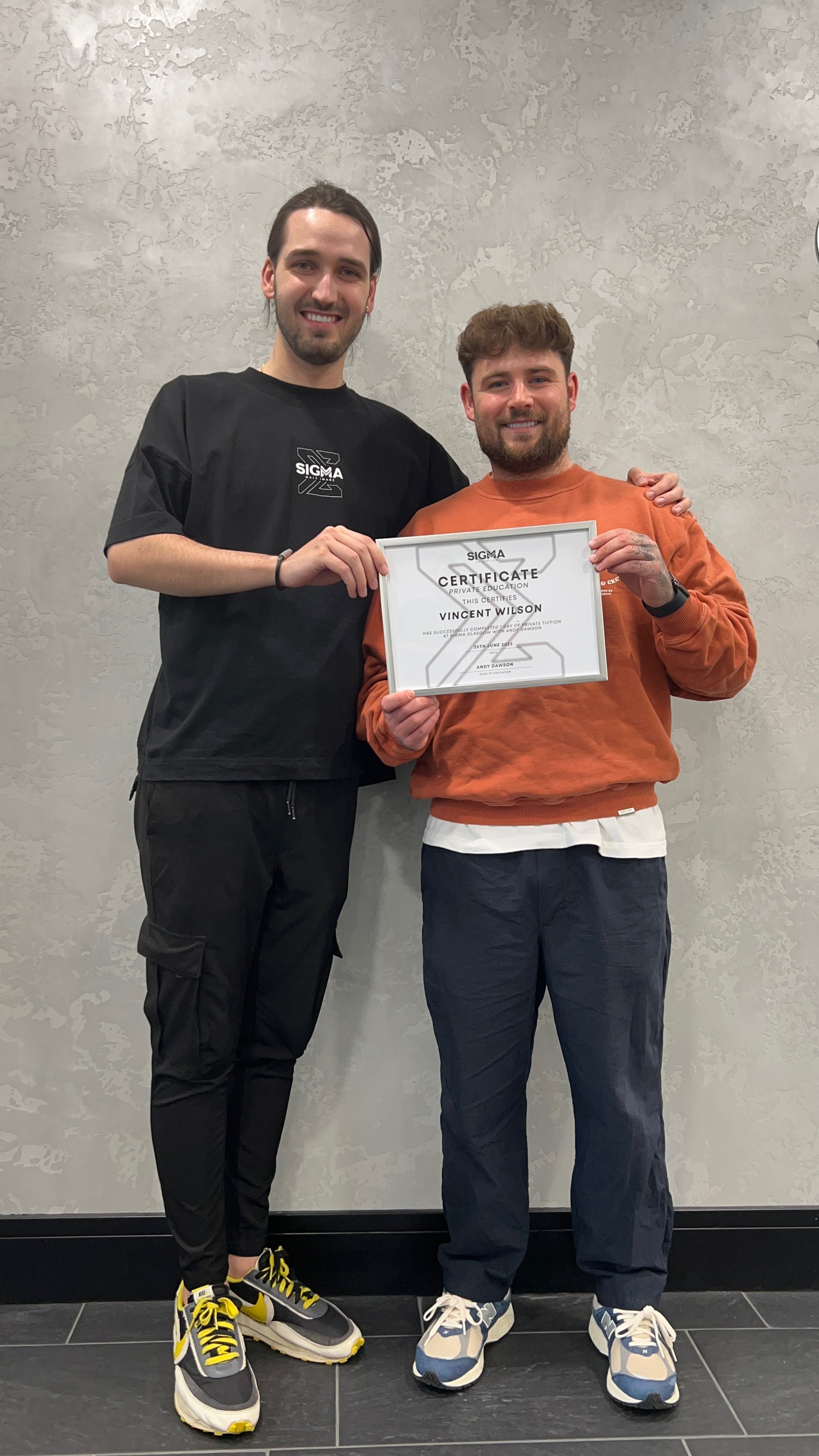 Two men standing together, smiling. The man on the right is holding a framed certificate that reads "Sigma Certificate of Private Education" awarded to Vincent Wilson. The man on the left is wearing a black Sigma t-shirt. They are standing against a 