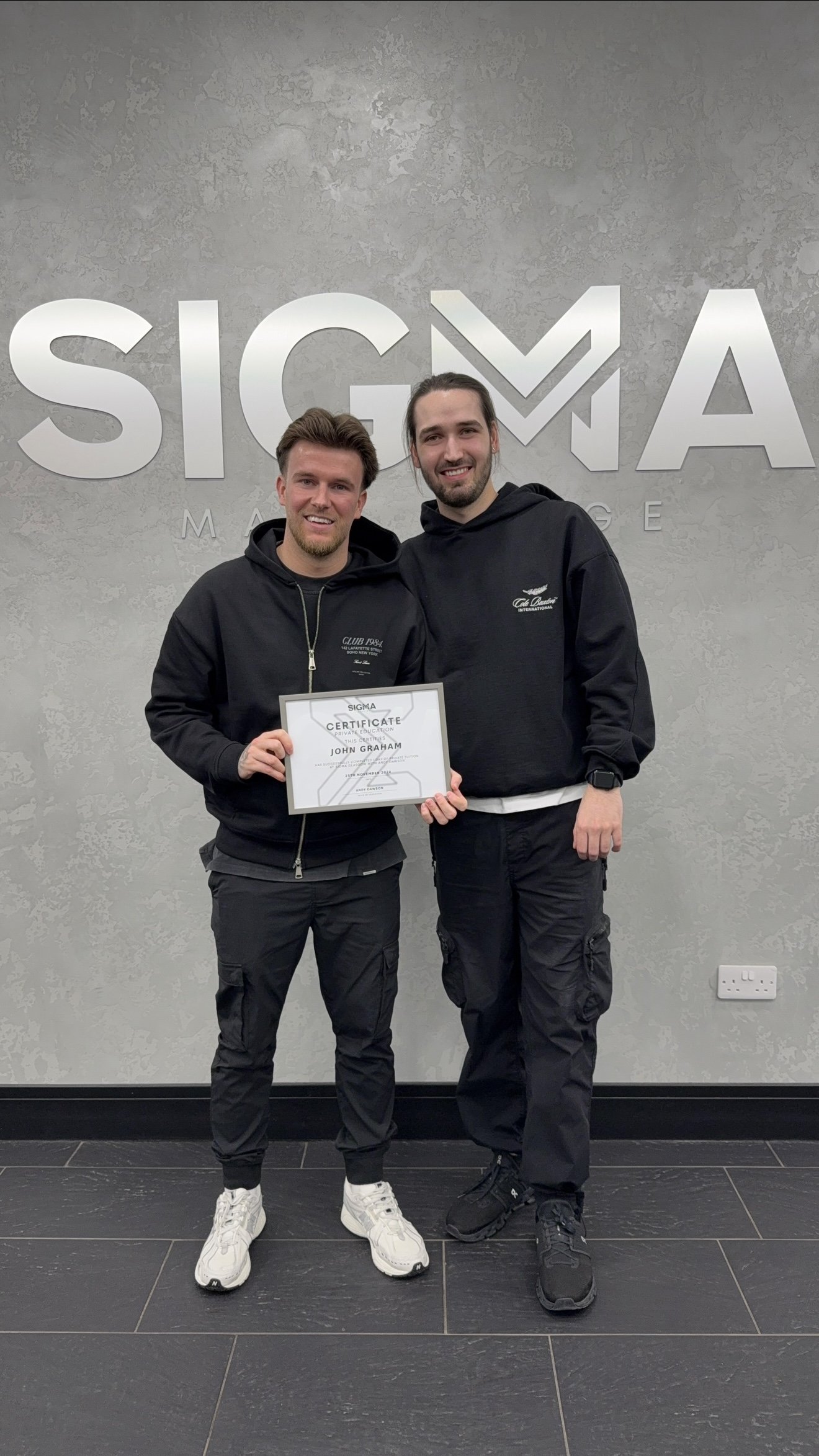 Two young men standing and smiling in front of a wall with a 'SIGMA' logo, one of them holding a certificate.
