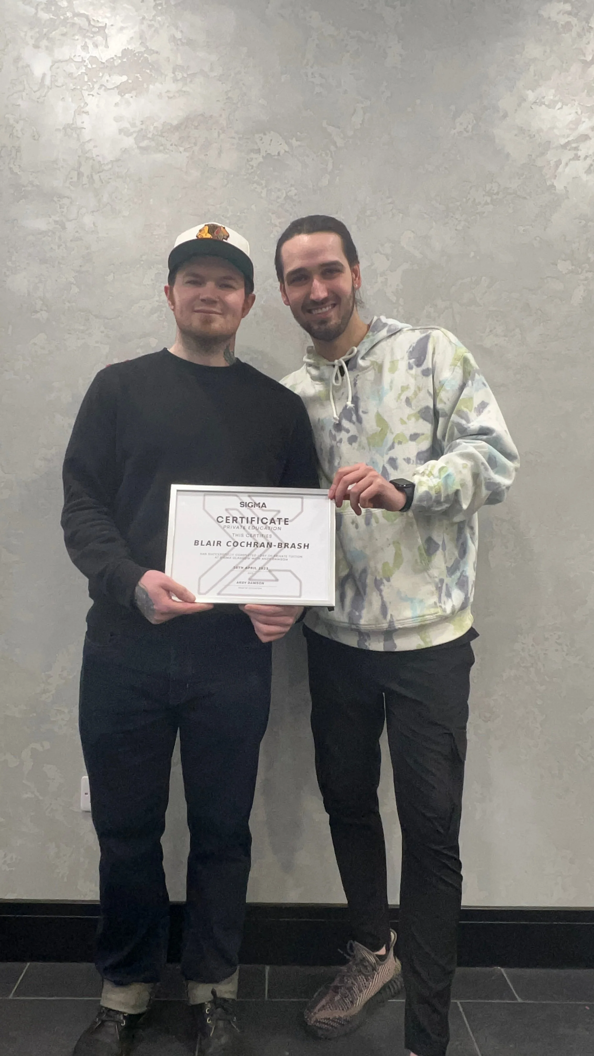 Two young men smiling, one holding a framed certificate that reads 'Sigma Certificate' with a name 'Blair Cochran-Brash'. They are standing against a textured gray wall.