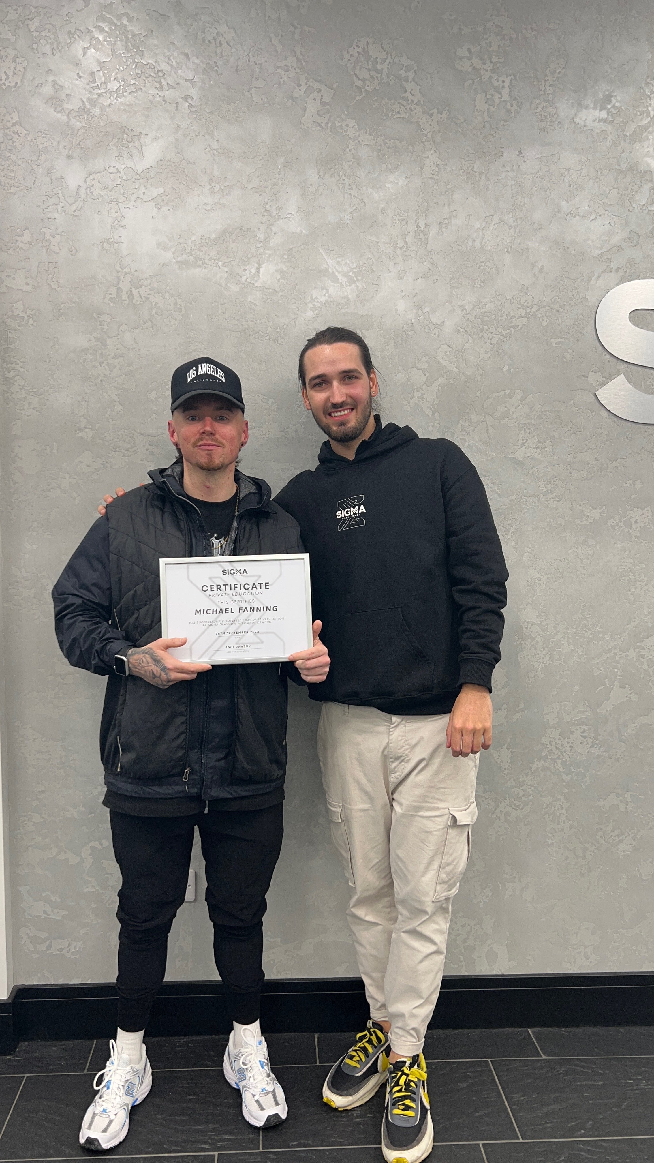 Two men standing side by side against a gray wall, one holding a certificate. The man on the left is wearing a black jacket, black pants, white sneakers, and a black baseball cap. The man on the right is in a black hoodie, beige pants, and black snea