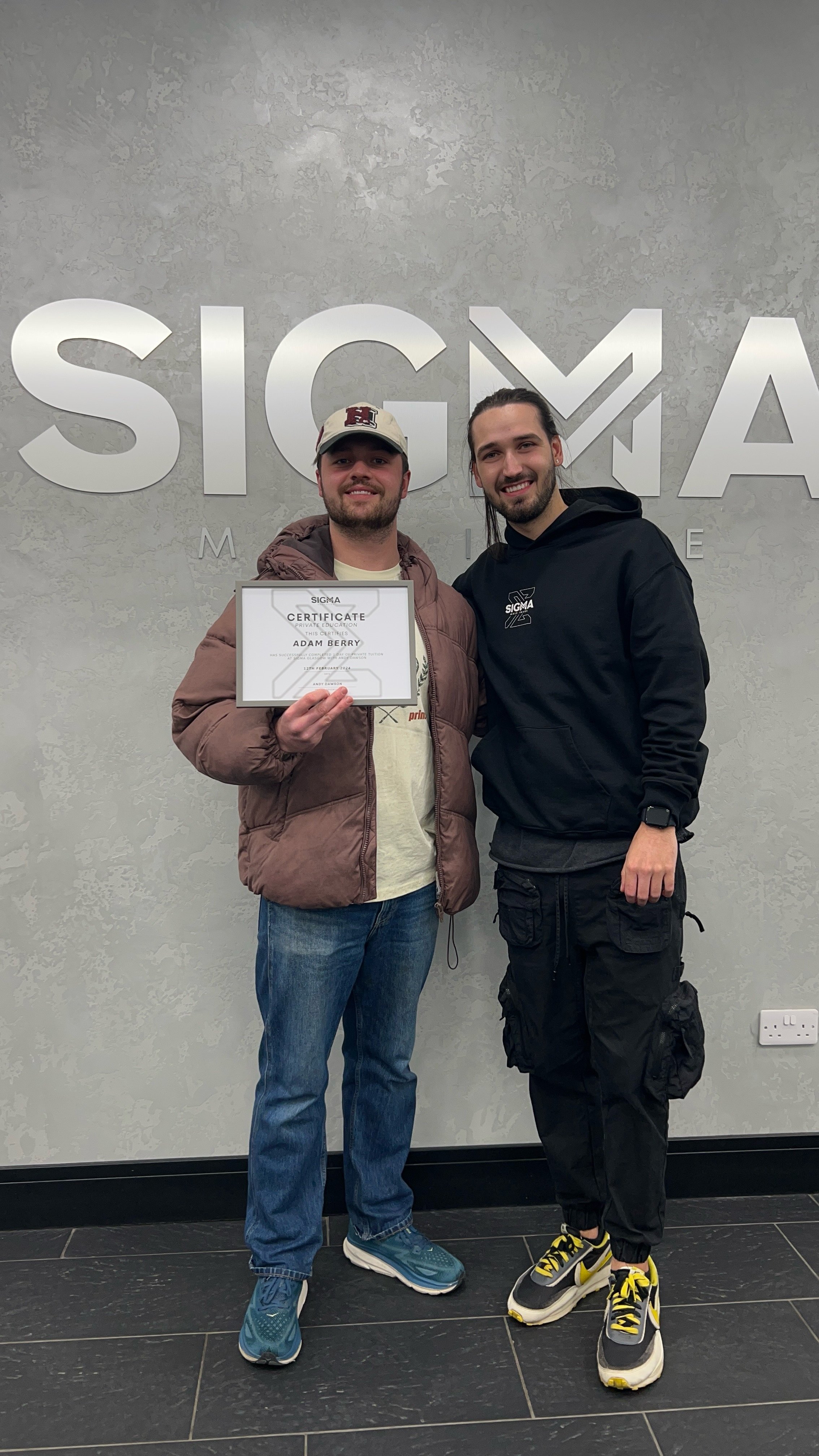 Two young men standing in front of a wall with a large Sigma logo, one holding a certificate, smiling for the camera.