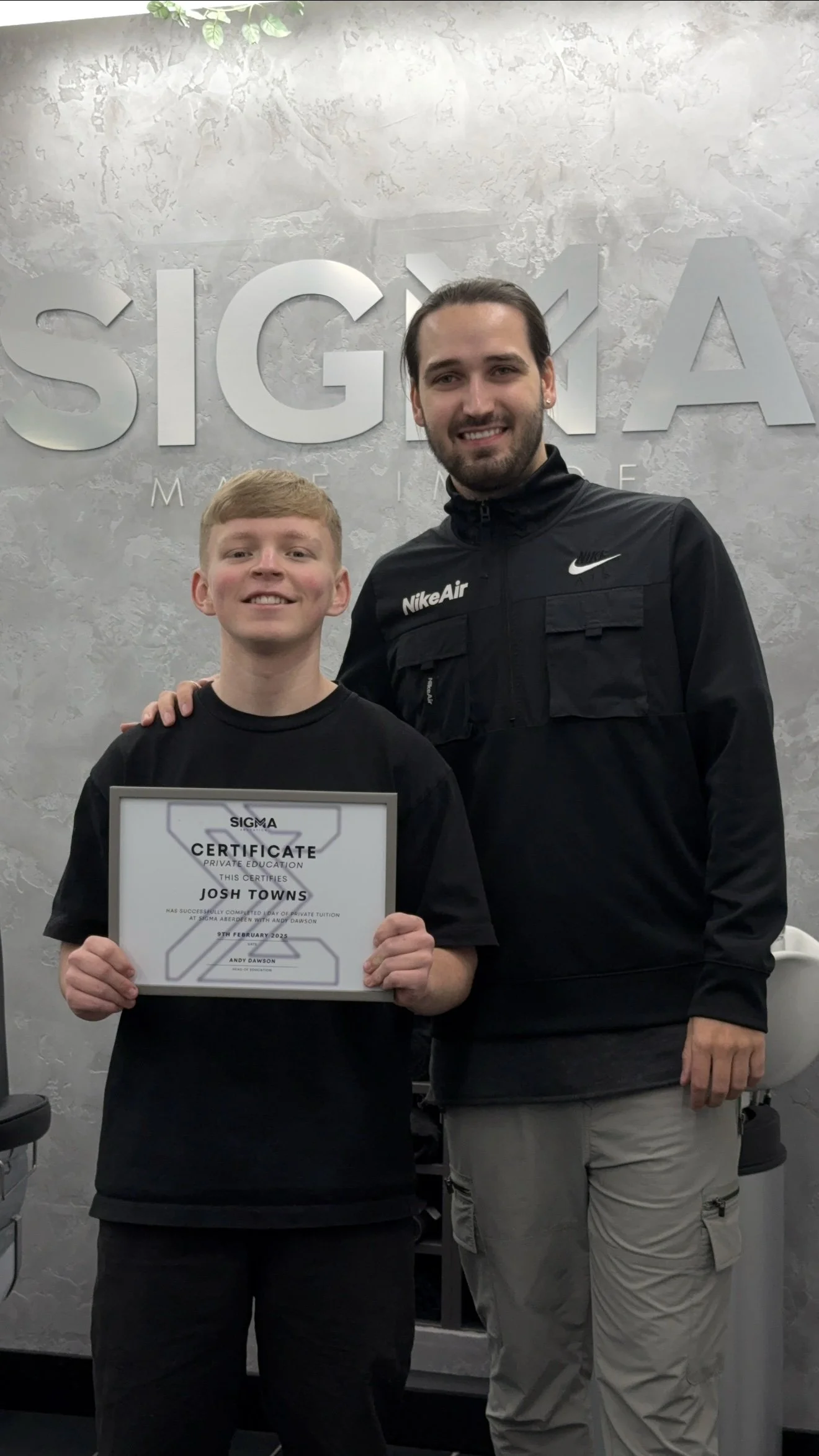 Two young men standing in front of a wall with the Sigma logo, one holding a certificate of completion at Sigma in front of the Sigma logo.