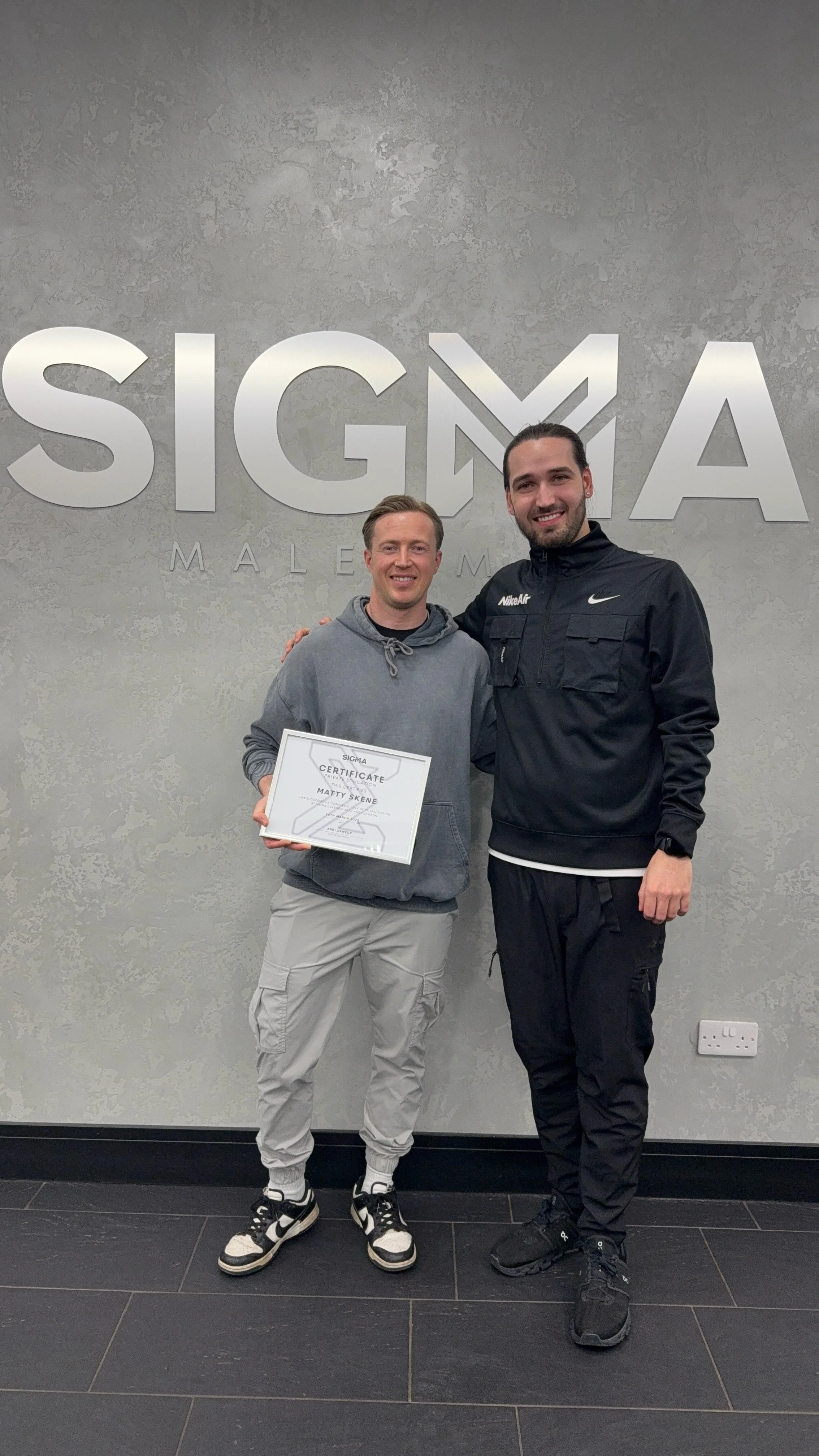 Two men standing in front of a wall with the Sigma logo, one holding a certificate. They are smiling and wearing casual and athletic clothing.