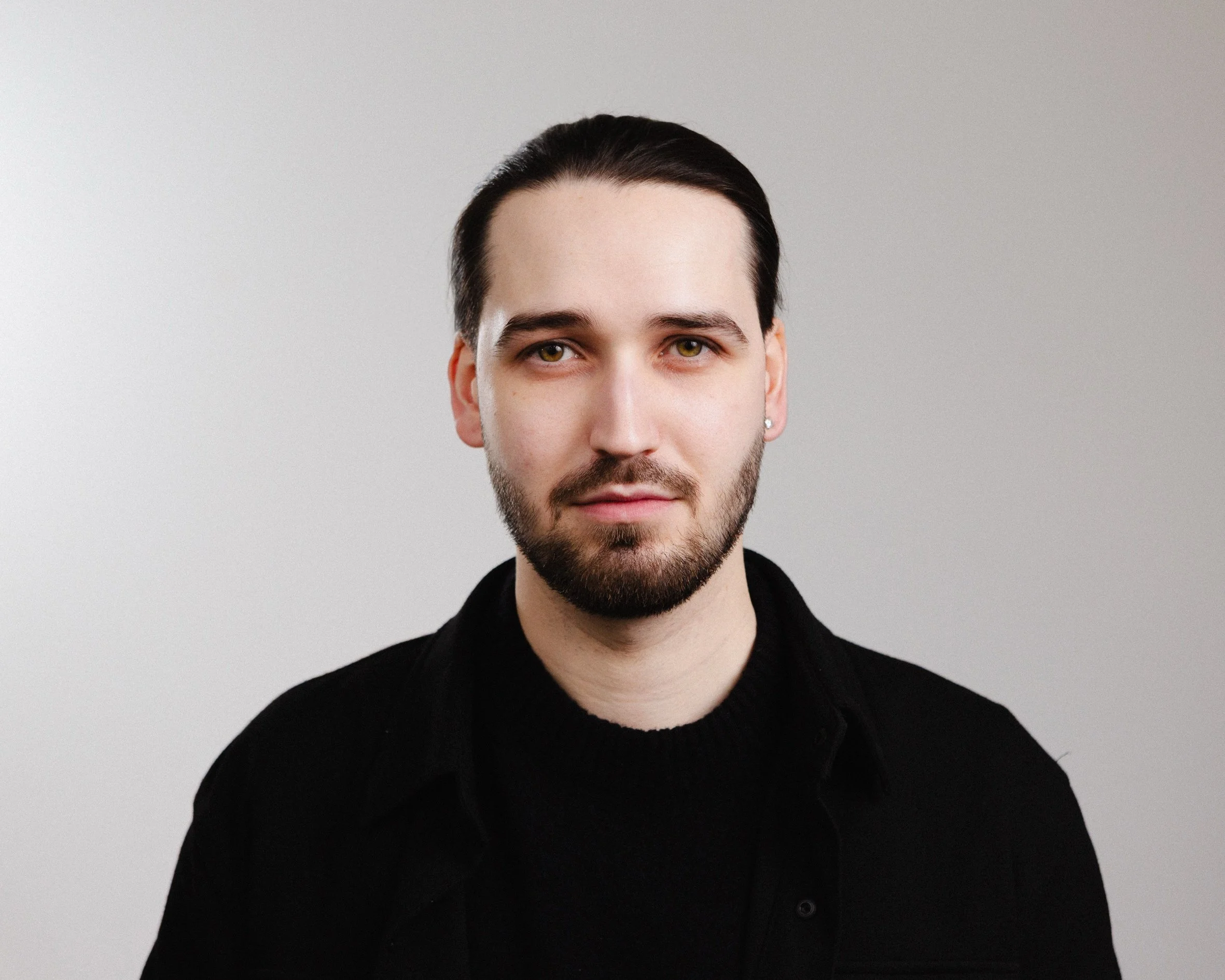 Portrait of a young man with dark hair and beard, wearing a black shirt, against a plain gray background.