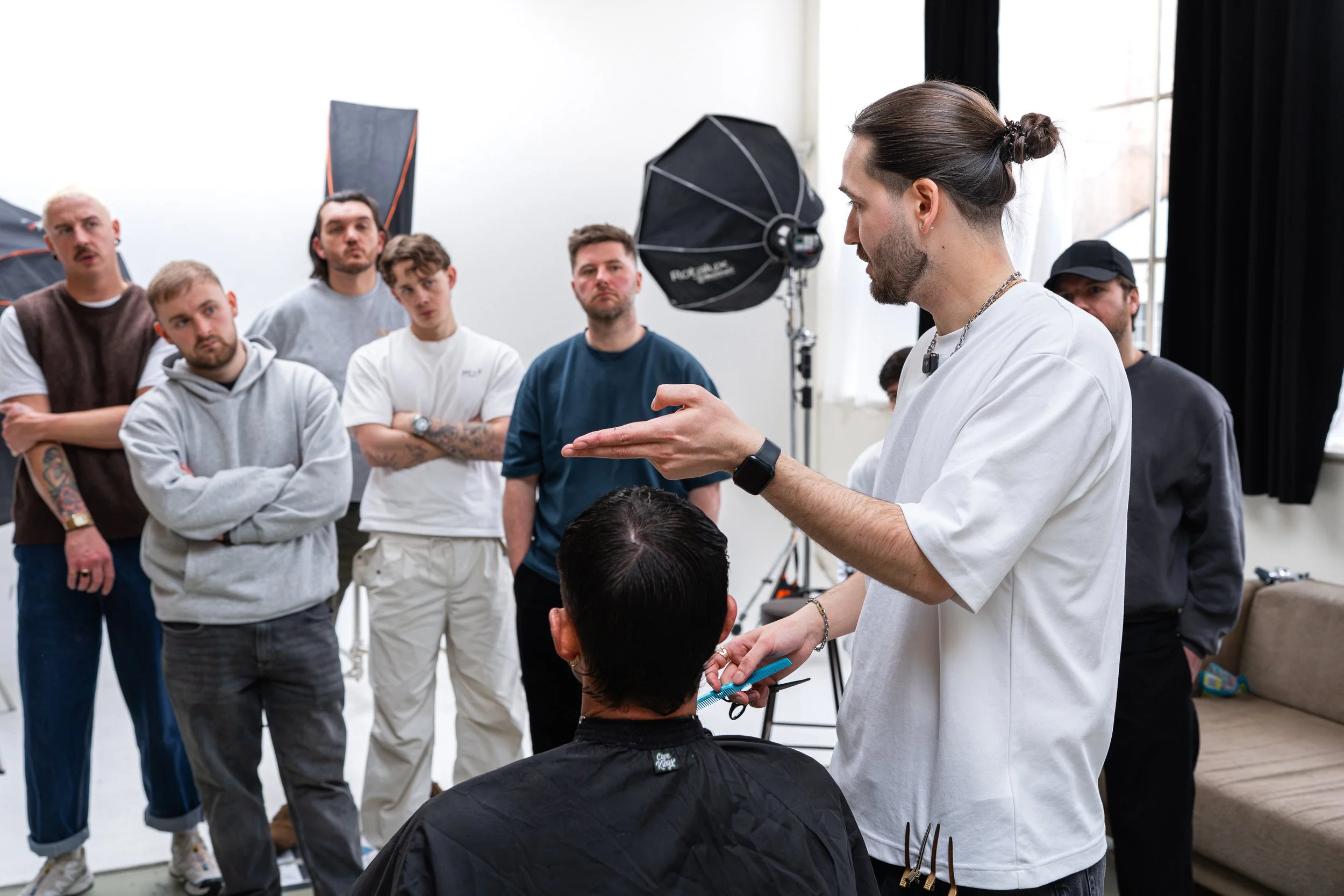 Male hairstylist giving a haircut demonstration to a group of men in a photography studio with lighting equipment and a couch.