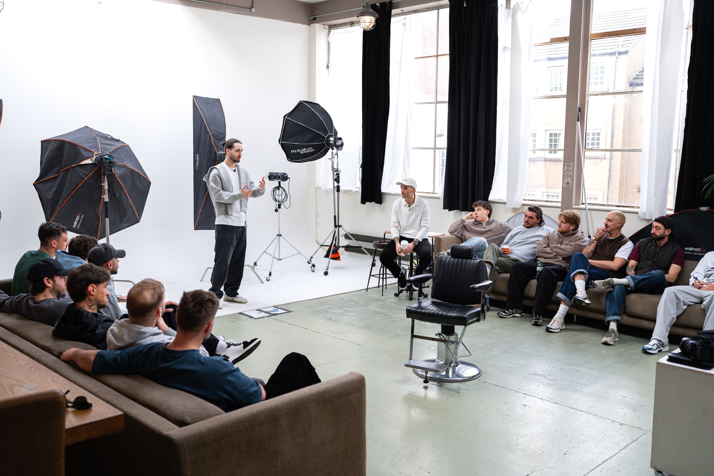 A filmmaker or instructor standing in front of a classroom of students during a video production or photography workshop in a spacious, well-lit studio with large windows, studio lighting equipment, and a white backdrop.