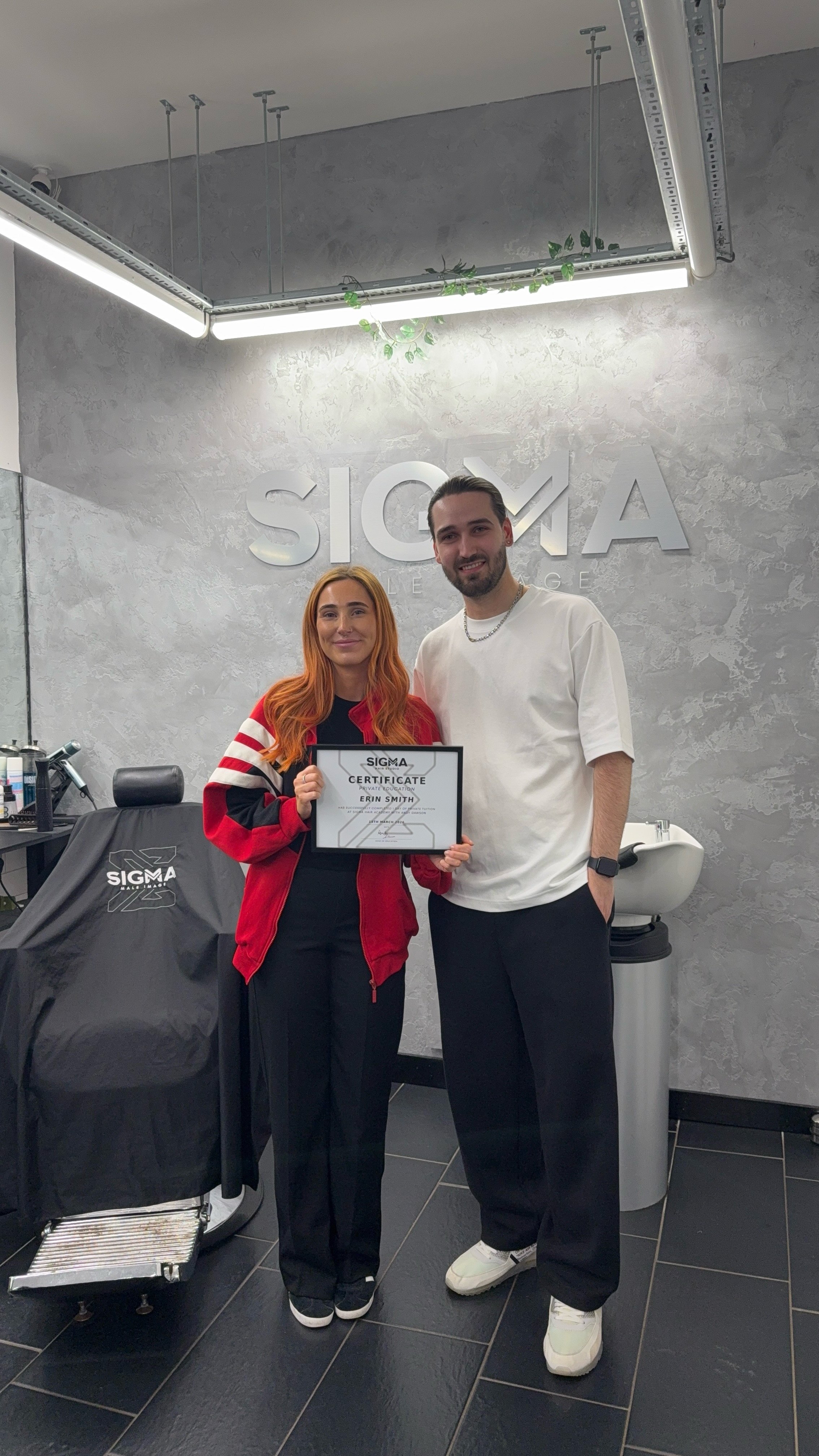 A woman with red hair and a man with dark hair and beard standing in a hair or beauty salon, holding a certificate. The salon has a sign that says "SIGMA" on the wall behind them, and the woman is wearing a red jacket with black pants while the man i