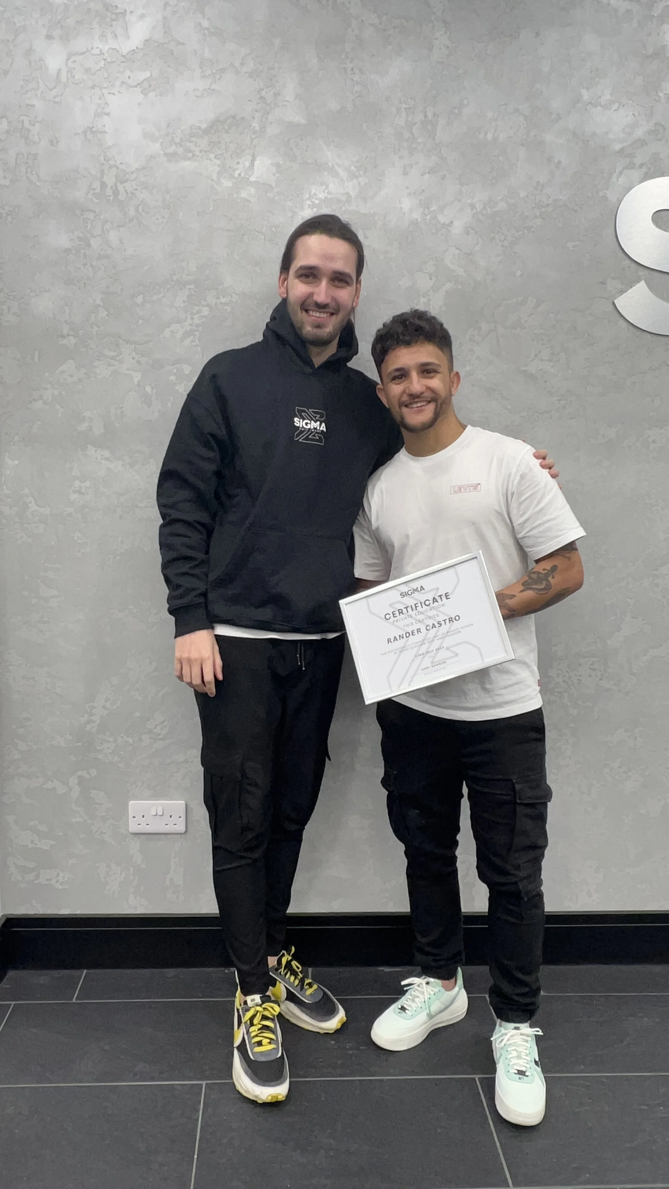 Two men standing indoors, posing for a photo. One man is taller, wearing a black Sigma hoodie and black pants with yellow and black sneakers. The shorter man is holding a certificate and wearing a white t-shirt, black pants, and white sneakers with t