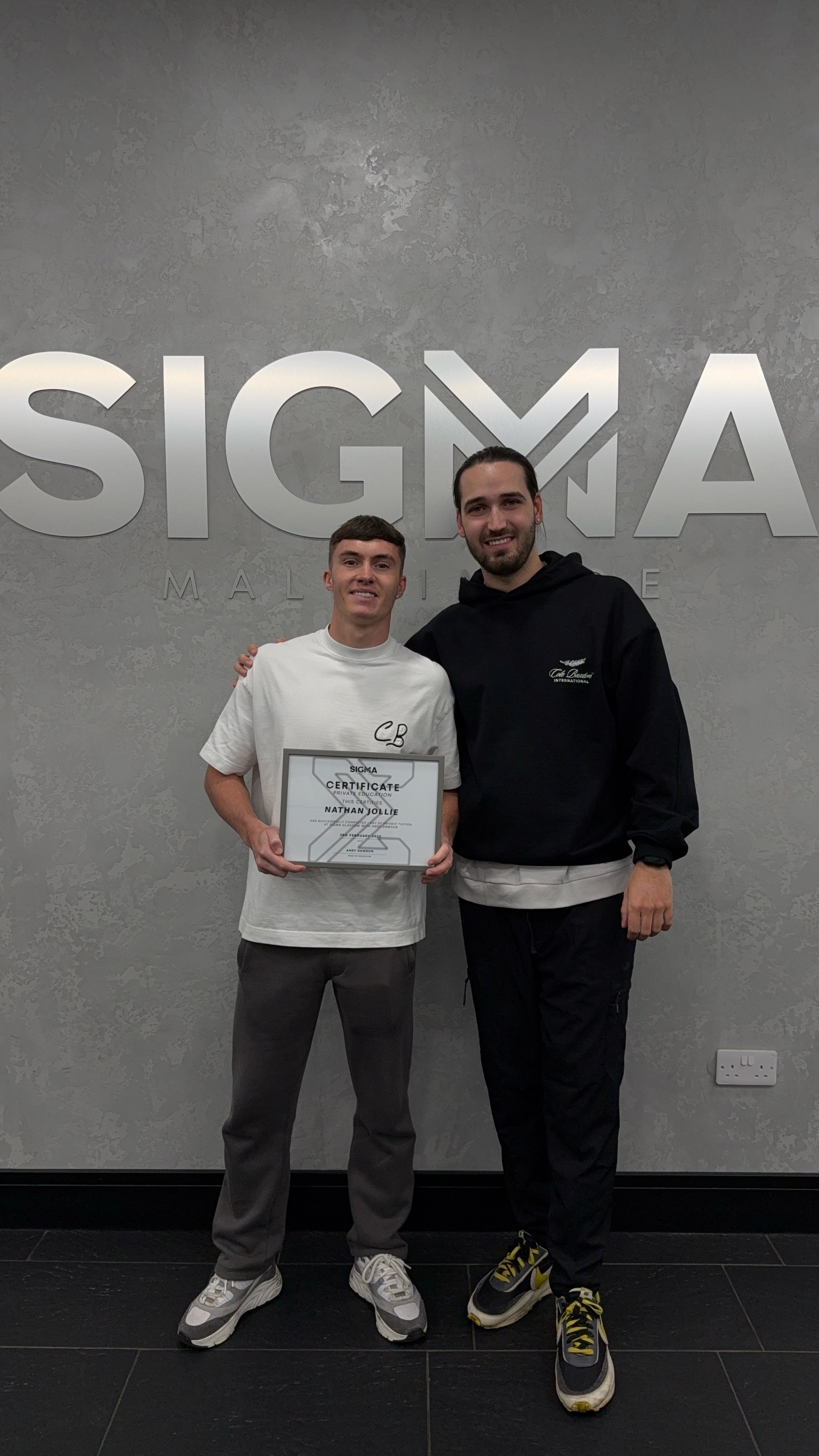Two men standing in front of a Sigma Maloka sign, one holding a certificate of achievement. The man on the left is wearing a white t-shirt with initials 'CB', gray pants, and sneakers, while the man on the right is wearing a black hoodie, black pants