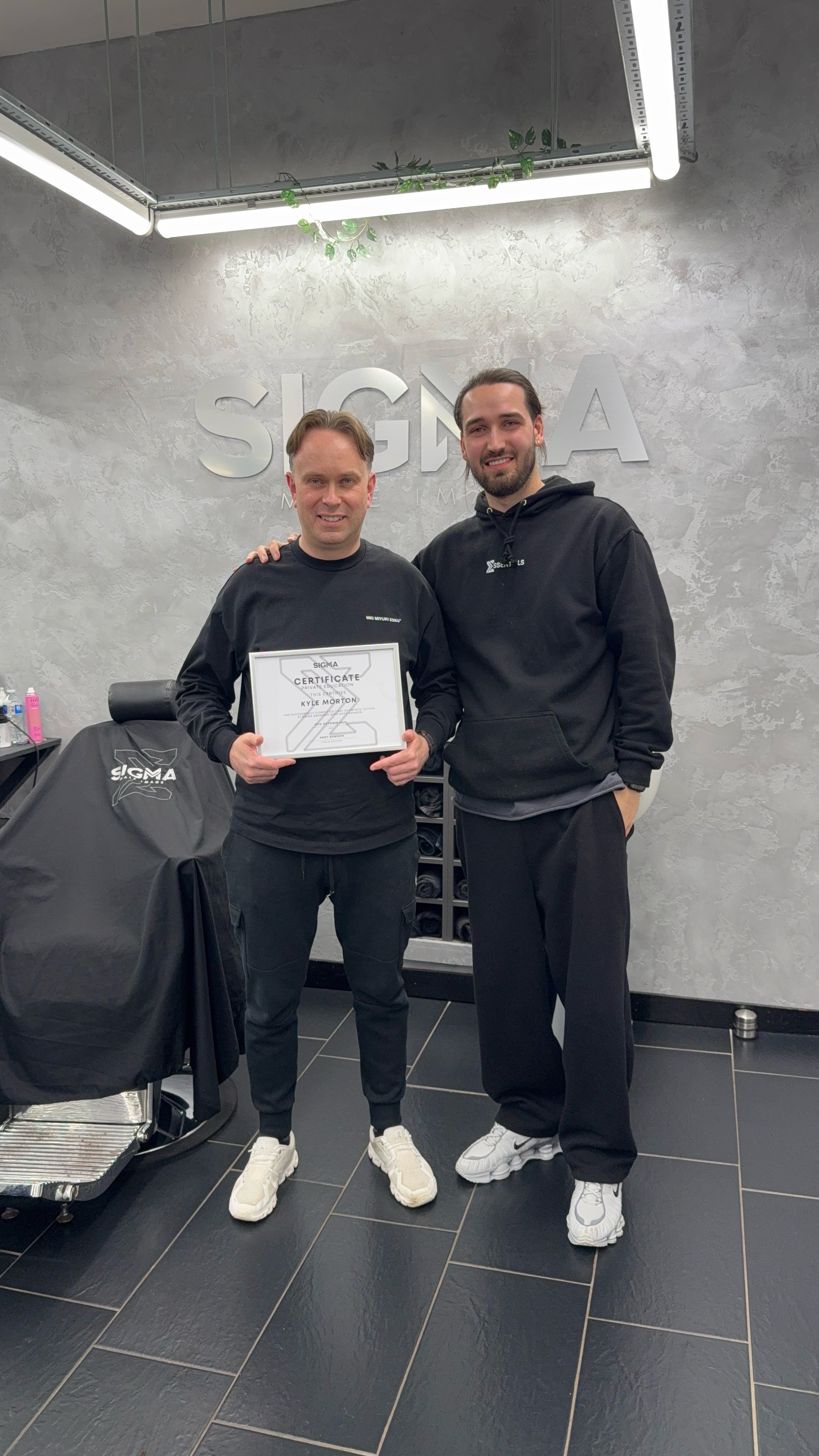 Two men standing together inside a modern gym or training studio, with one holding a certificate. Both are dressed in black athletic clothing and white sneakers. The background features a textured grey wall with the word 'SIGMA' mounted on it, and fi