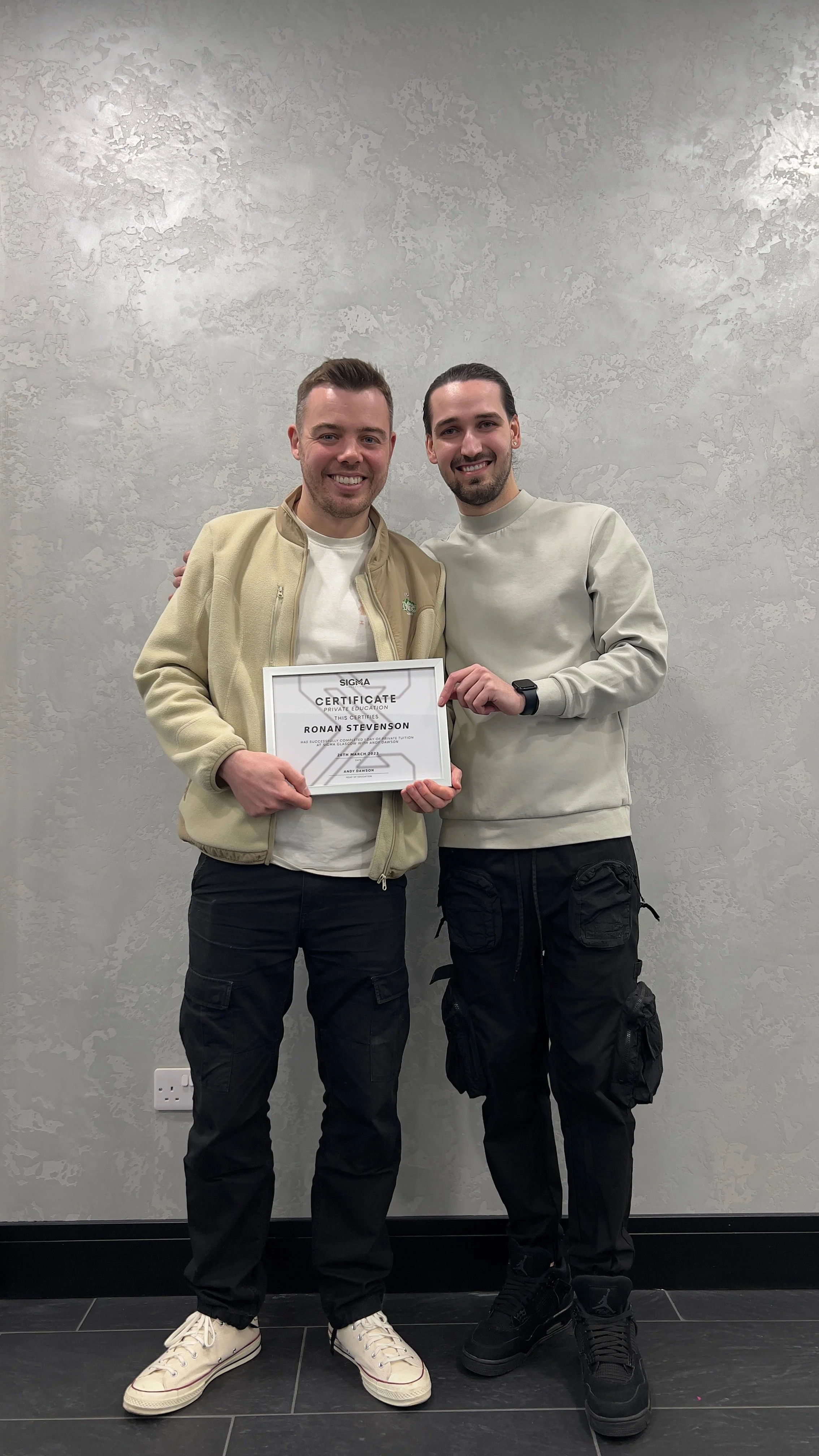 Two young men standing side by side, smiling, one holding a framed certificate. The man on the left is wearing a beige jacket, white shirt, black cargo pants, and white sneakers. The man on the right is wearing a light gray sweatshirt, black cargo pa