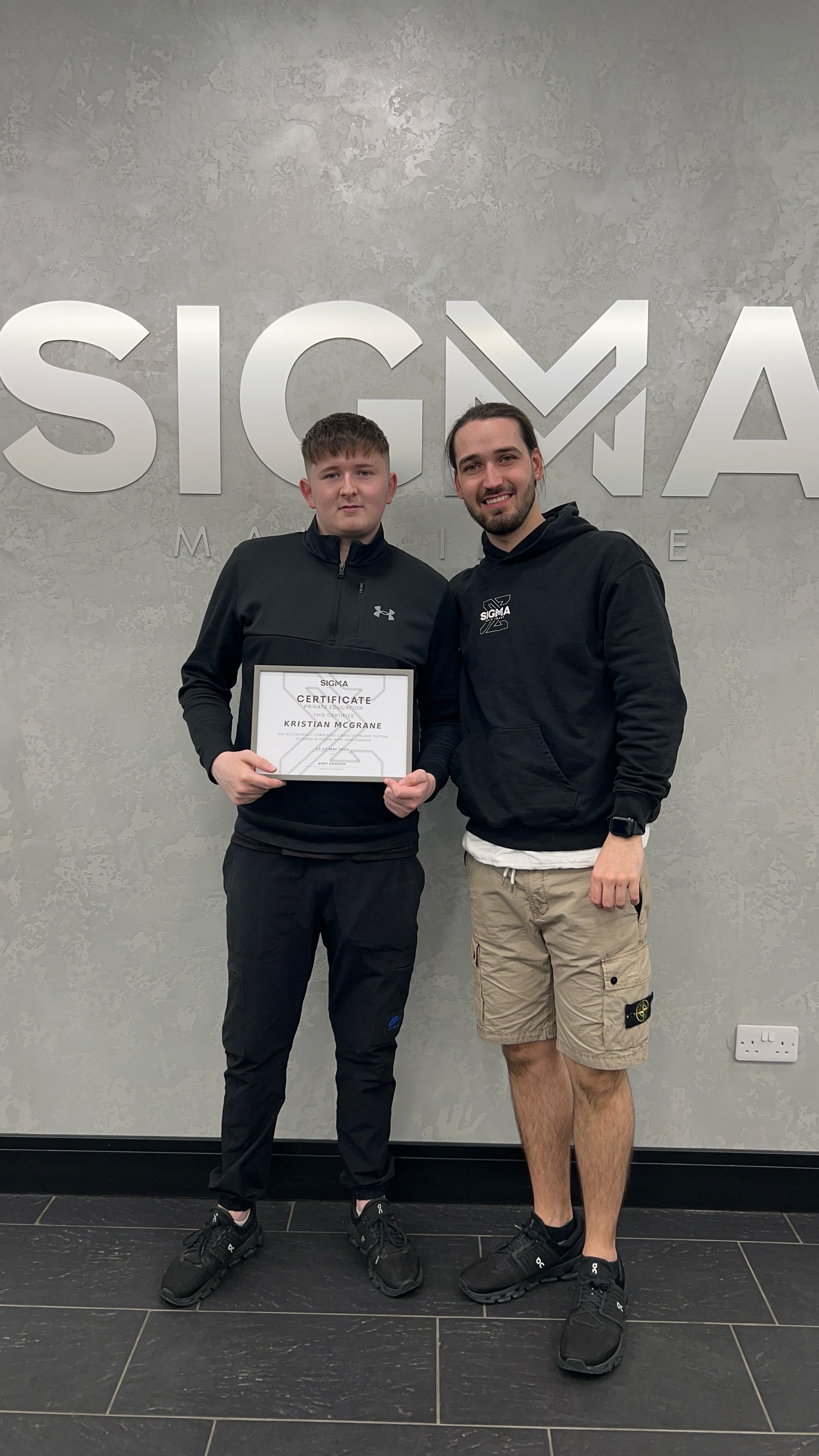Two men standing in front of a wall with the SIGMA logo, one is holding a certificate. The man on the left has short brown hair, is wearing a black jacket, black pants, and black shoes. The man on the right has dark hair tied back, a beard, and is we