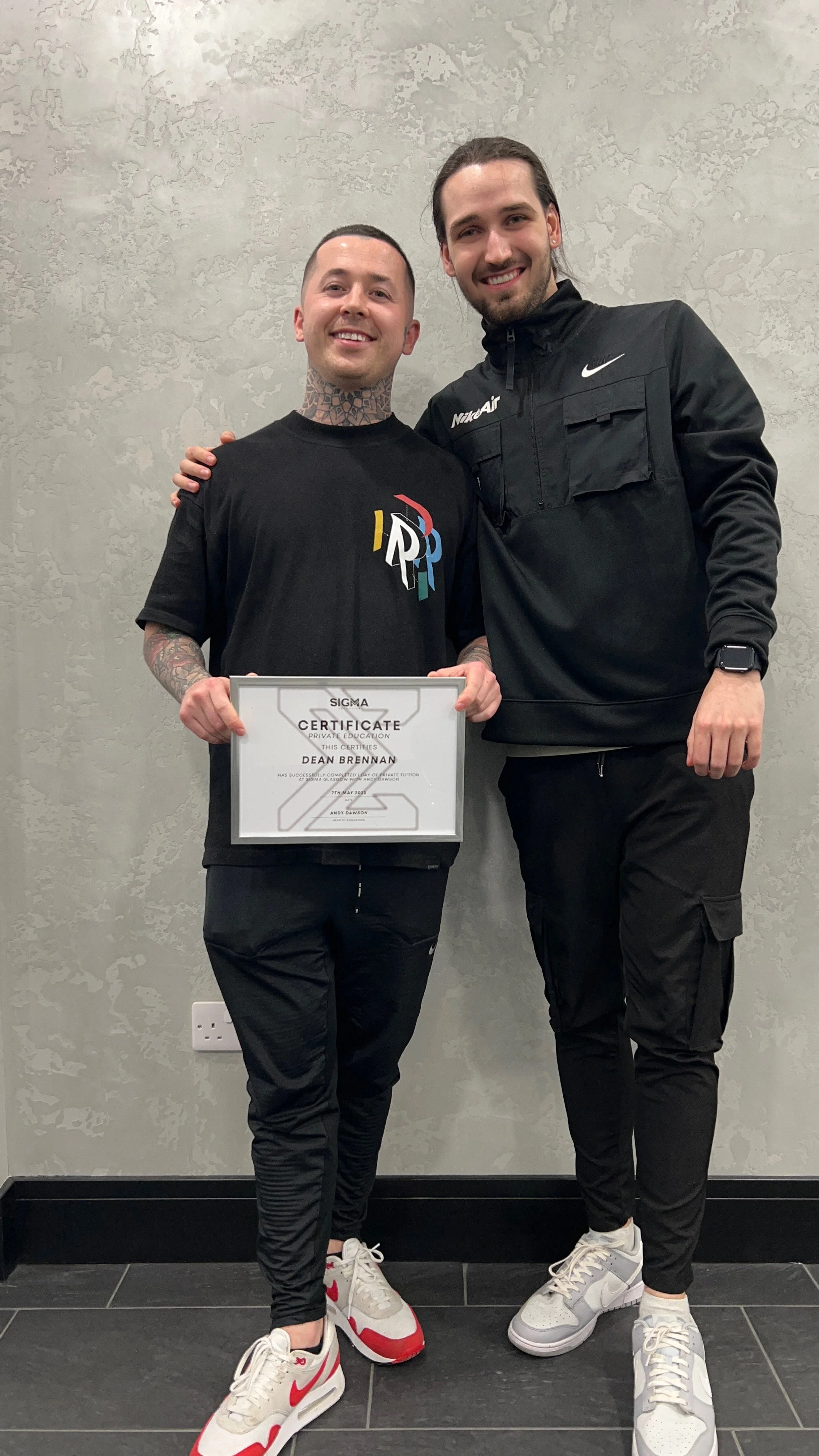 Two young men standing side by side, one holding a framed certificate. Both are smiling and wearing athletic clothing. The man on the left has tattoos on his neck and arms, short dark hair, and is wearing a black t-shirt with a colorful logo. They ar