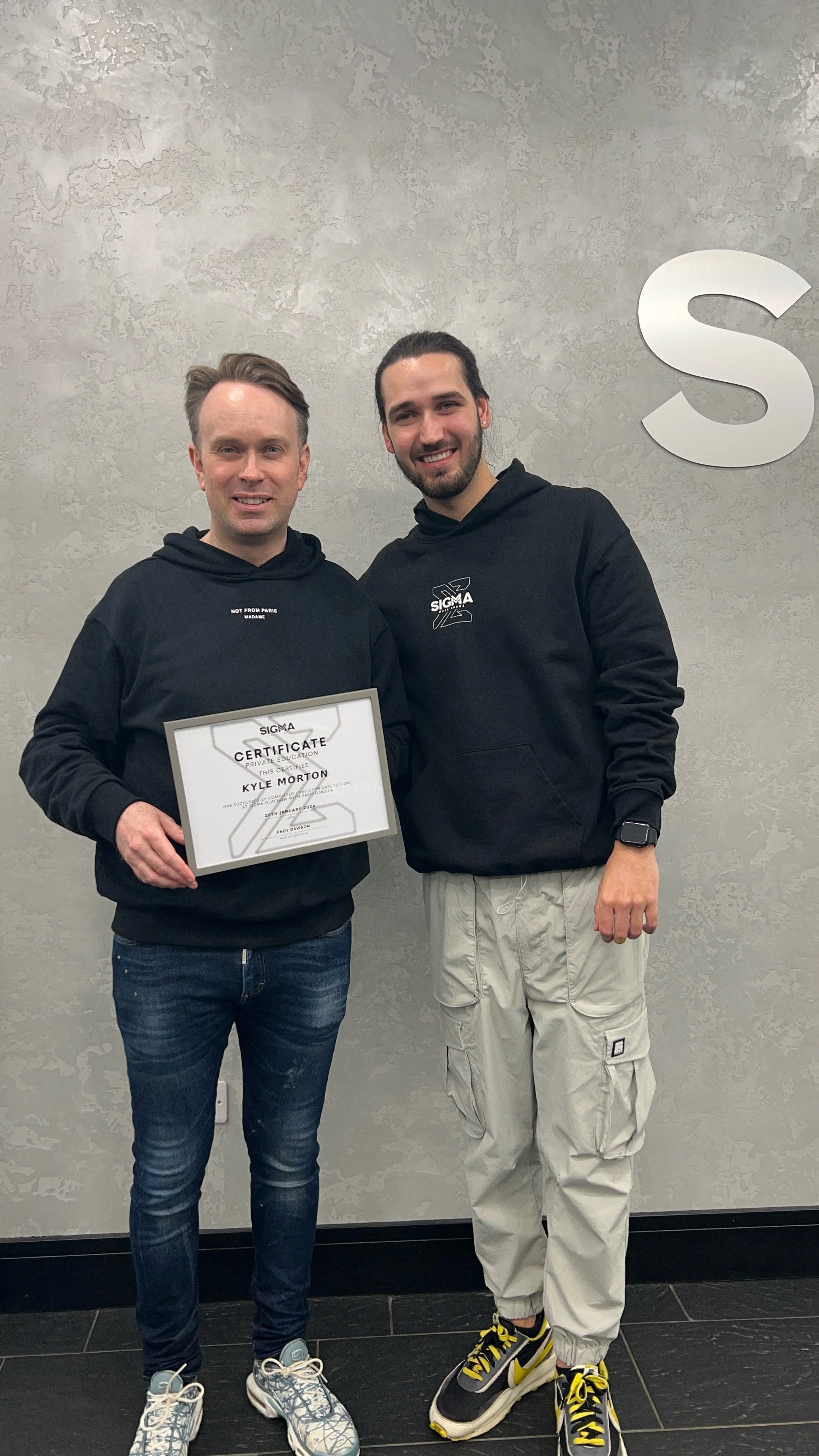 Two men standing indoors, one holding a Sigma certificate, both wearing black Sigma hoodies, smiling, with a large Sigma letter on the wall behind them.