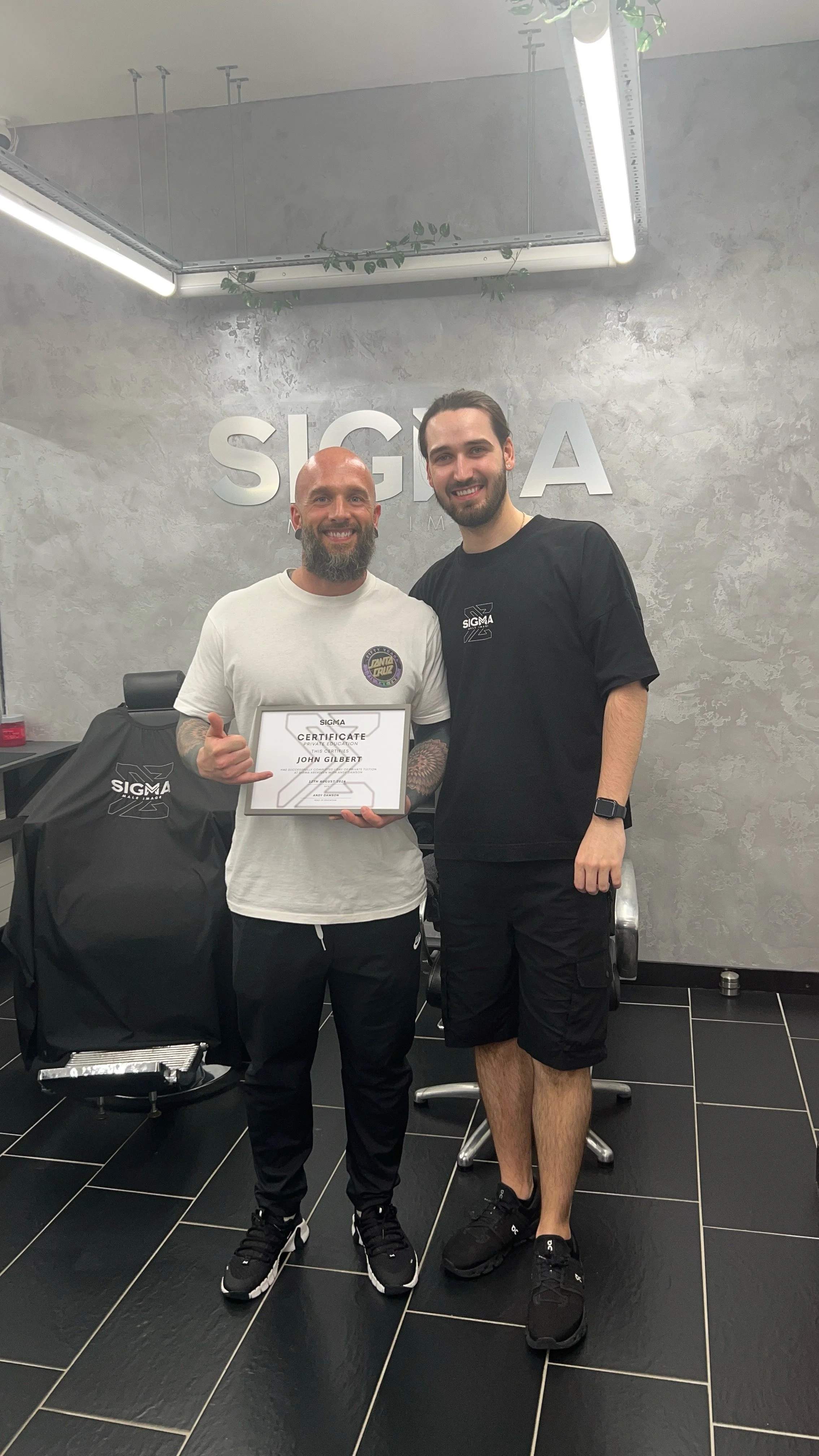 Two men standing inside a fitness or gym facility, smiling for the camera. The man on the left is holding a certificate and making a shaka sign with his right hand; he has a beard and tattoos on his arms. The man on the right is taller, has dark hair