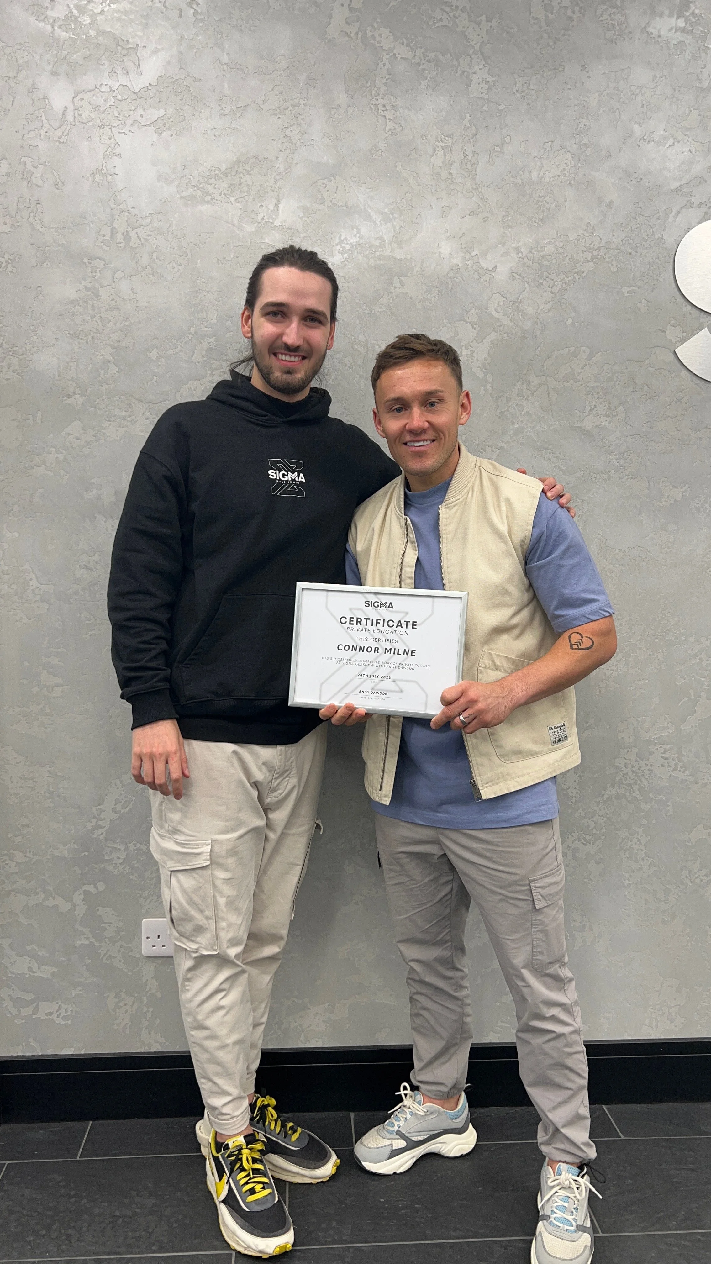 Two young men standing side by side, smiling, with one holding a certificate of achievement. The background is a textured gray wall.