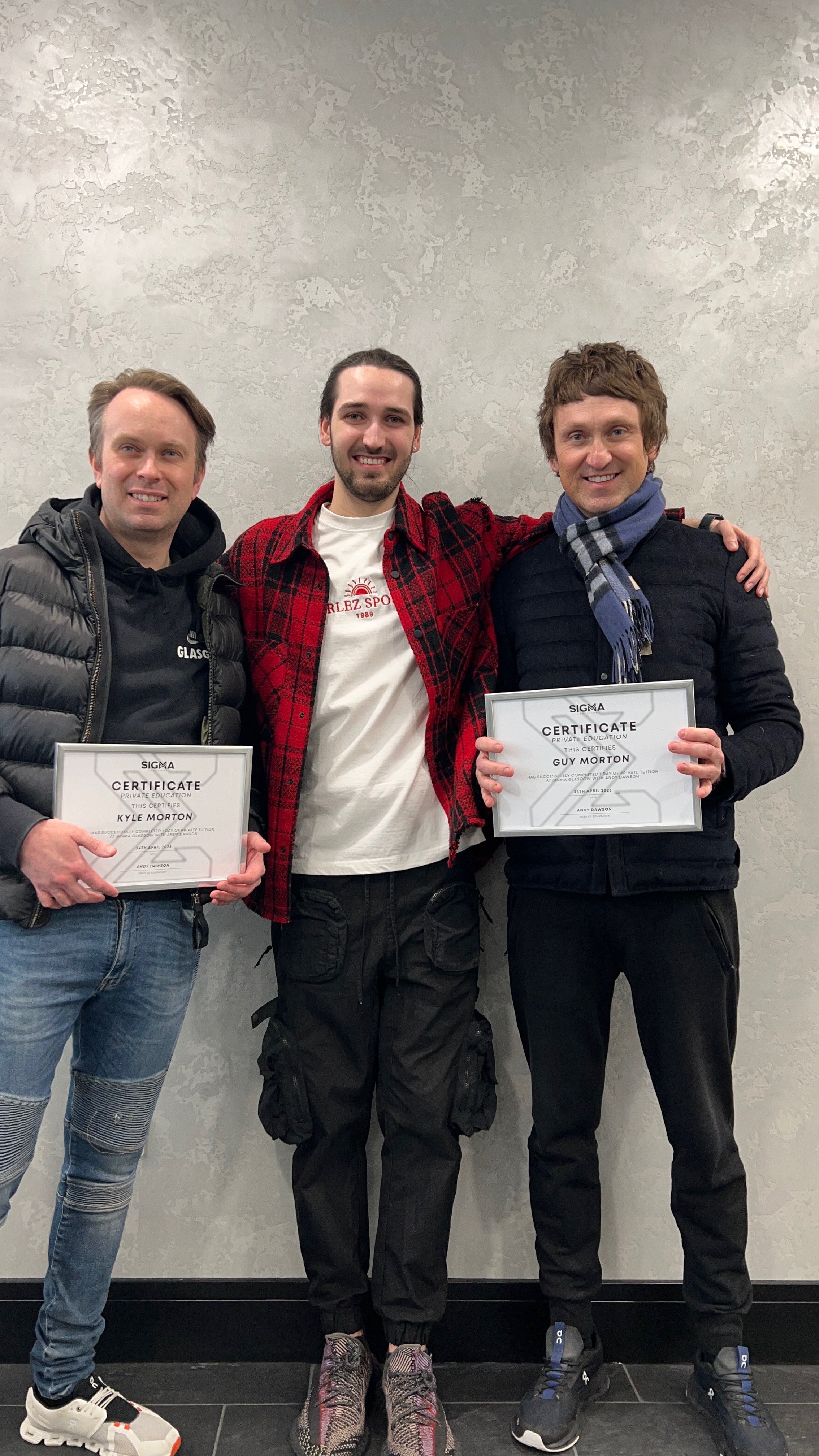 Three young men standing together, two holding certificates, smiling, in front of a gray textured wall.