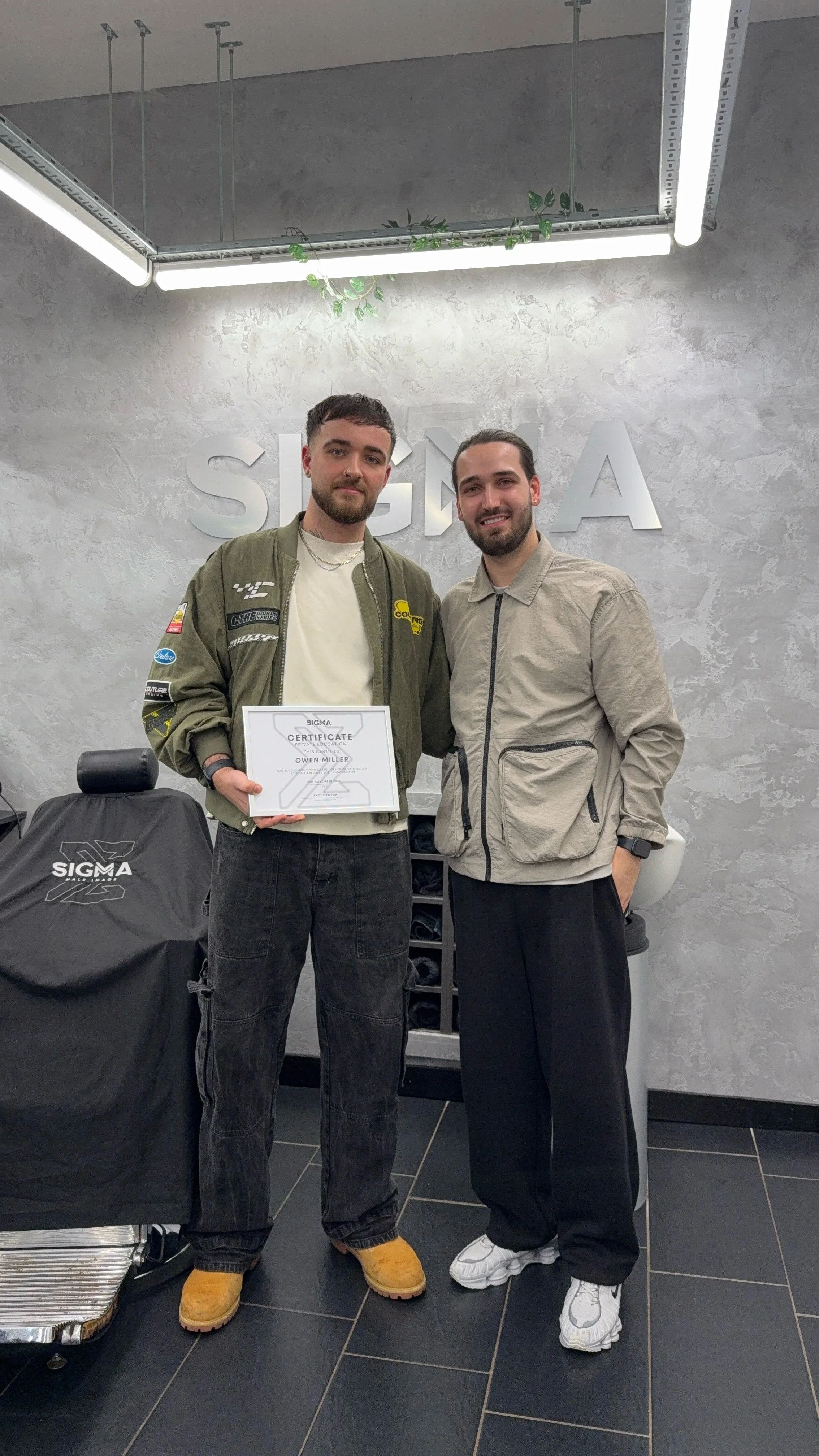 Two young men standing in a room with a gray textured wall, one holding a certificate, both smiling. Behind them is a sign that reads 'SIGMA,' and to the left, a covered object with the SIGMA logo. The room features modern lighting and minimal decor.