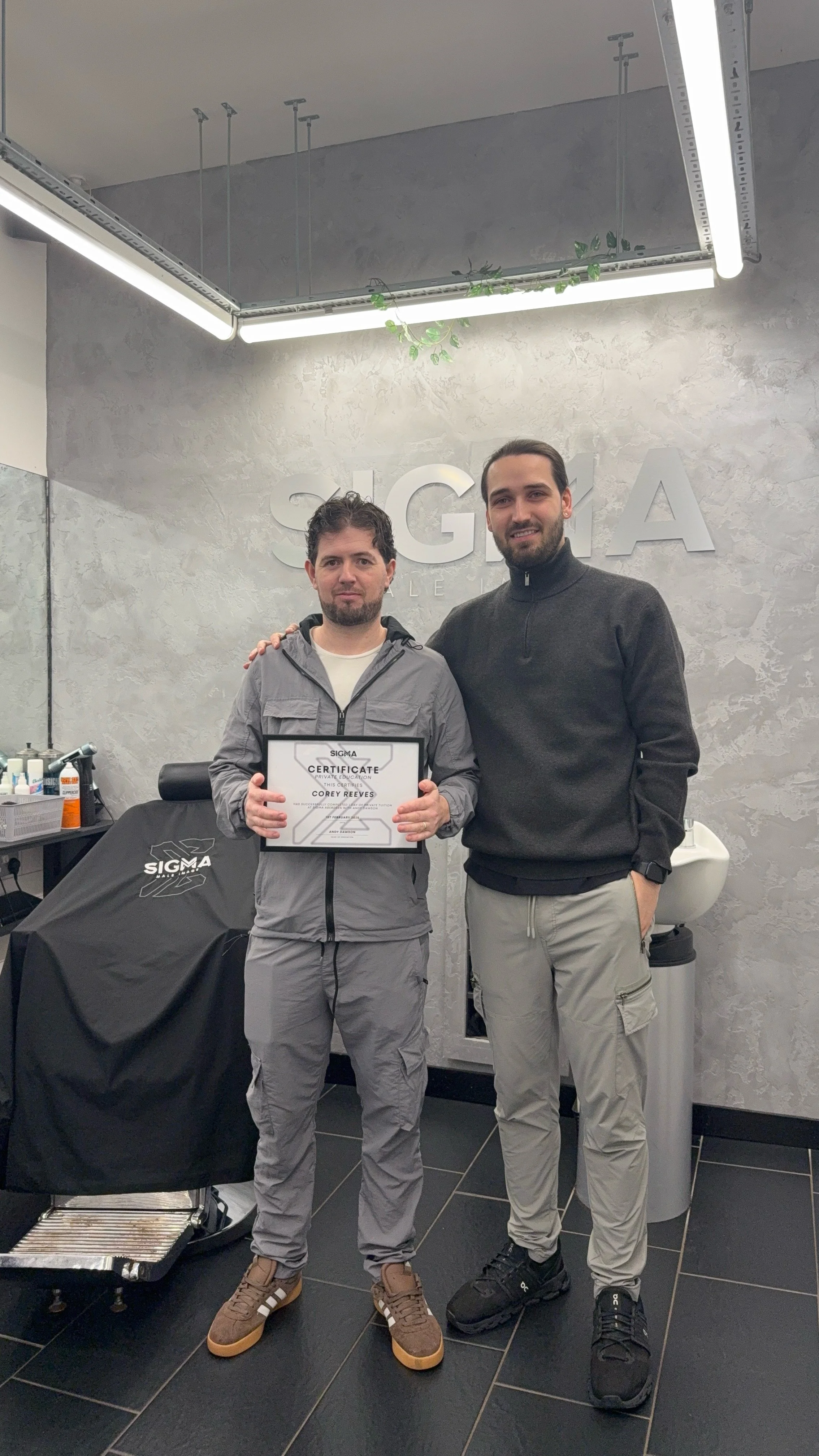 Two men standing indoors, one holding a certificate, in front of a wall with the word SIGMA. One man is wearing a grey jacket and pants, the other is in a black jacket and light grey pants. They are smiling and posing for the photo.