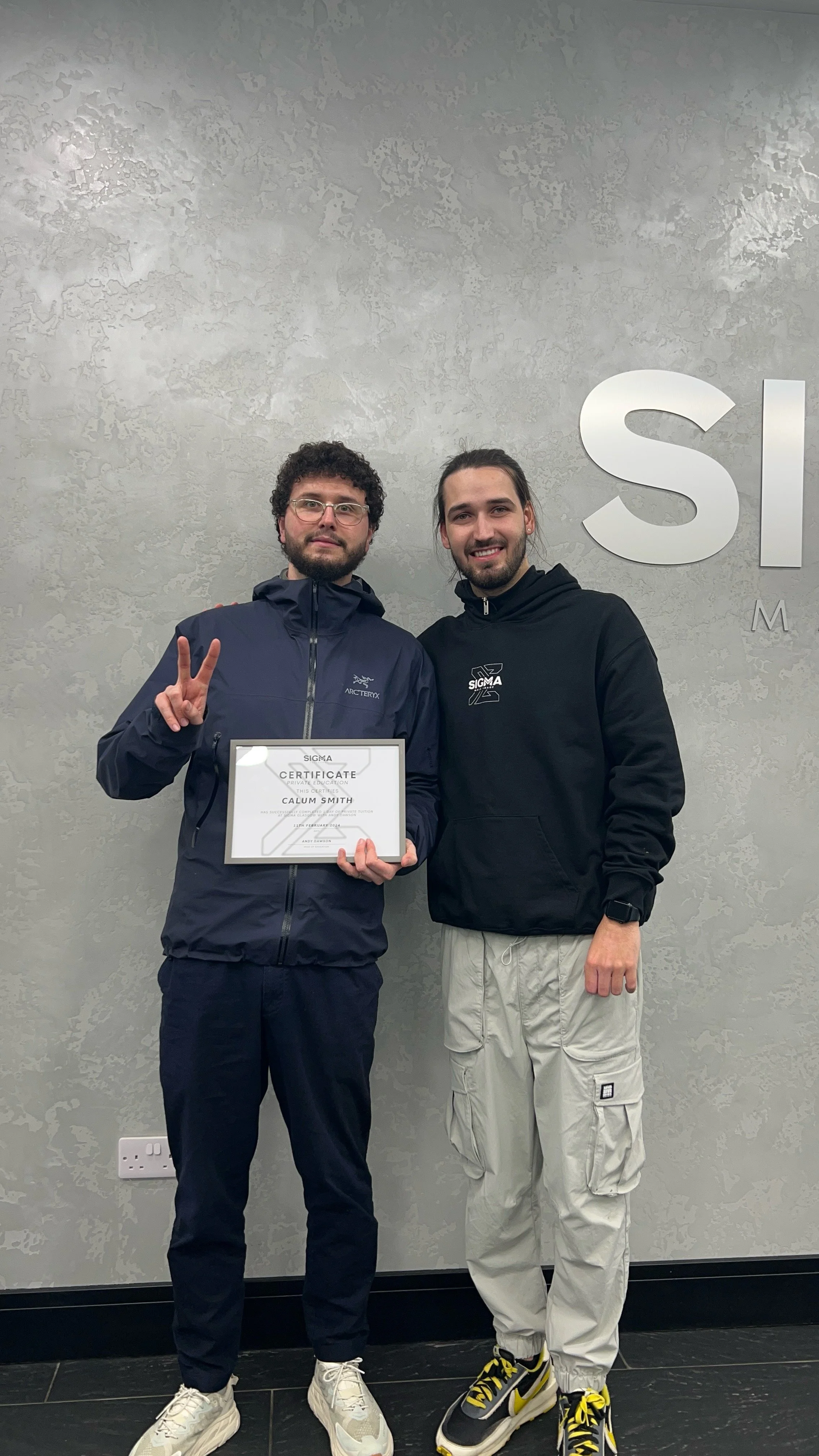Two young men standing in front of a grey wall, one holding a certificate, and the other flashing a peace sign. The wall has the word 'SI' visible in large letters.