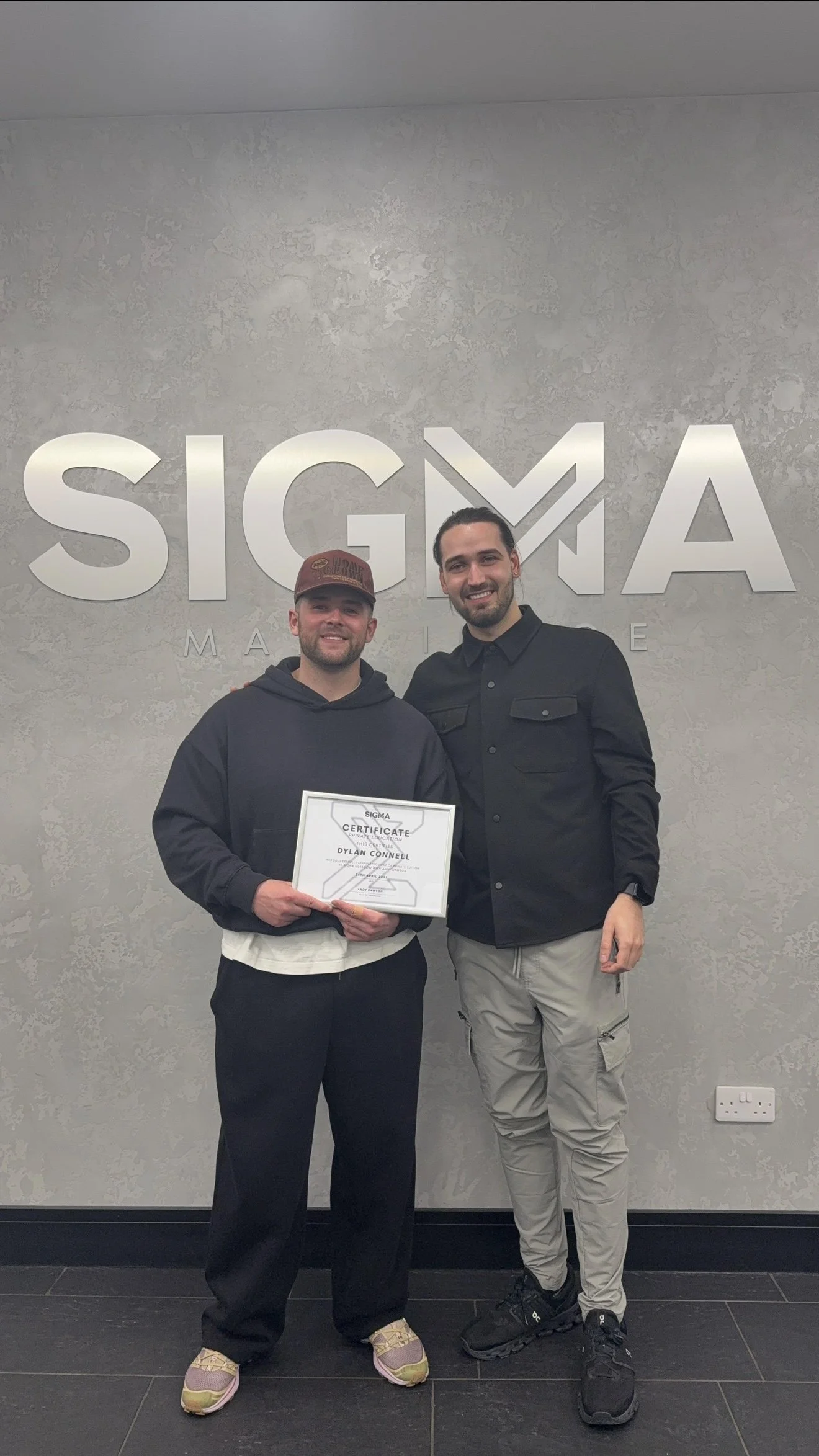 Two men standing in front of a wall with 'SIGMA' letters. The man on the left is holding a certificate. Both are smiling.
