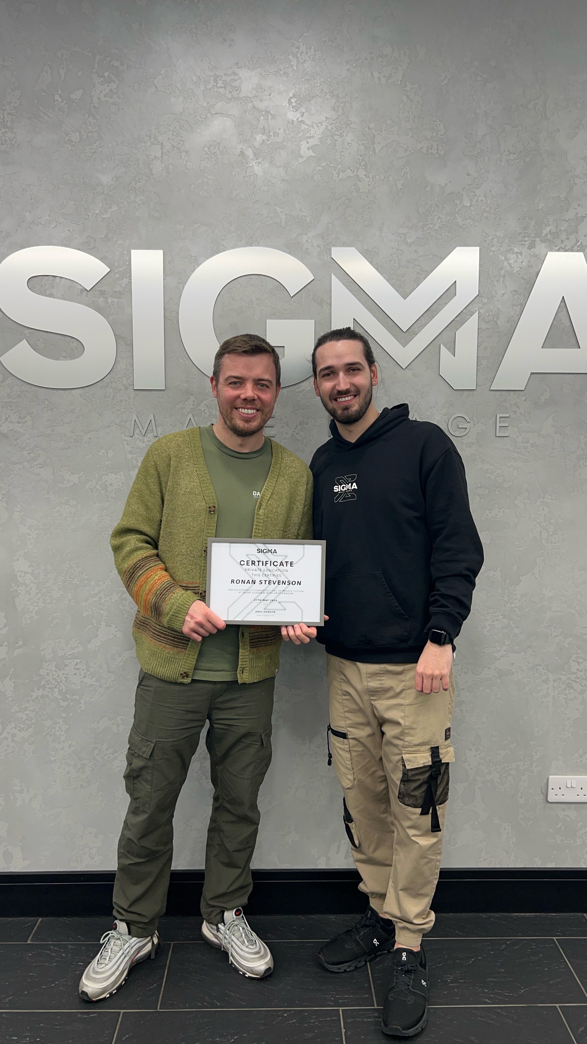 Two young men smiling, holding a certificate between them, standing in front of a wall with the Sigma logo, celebrating an achievement at Sigma.