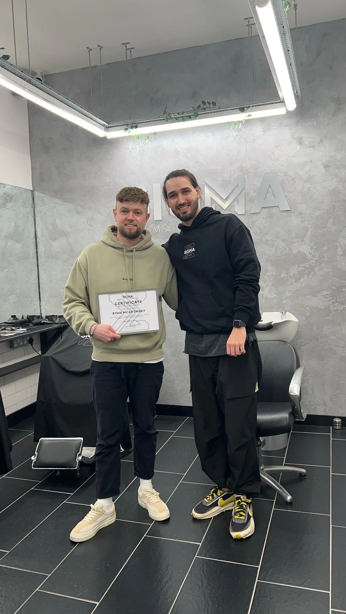 Two men standing in a modern barbershop while one holds a certificate. The man on the left wears a beige hoodie, black pants, and beige sneakers. The man on the right wears a black hoodie, black cargo pants, and black sneakers with yellow accents. Th