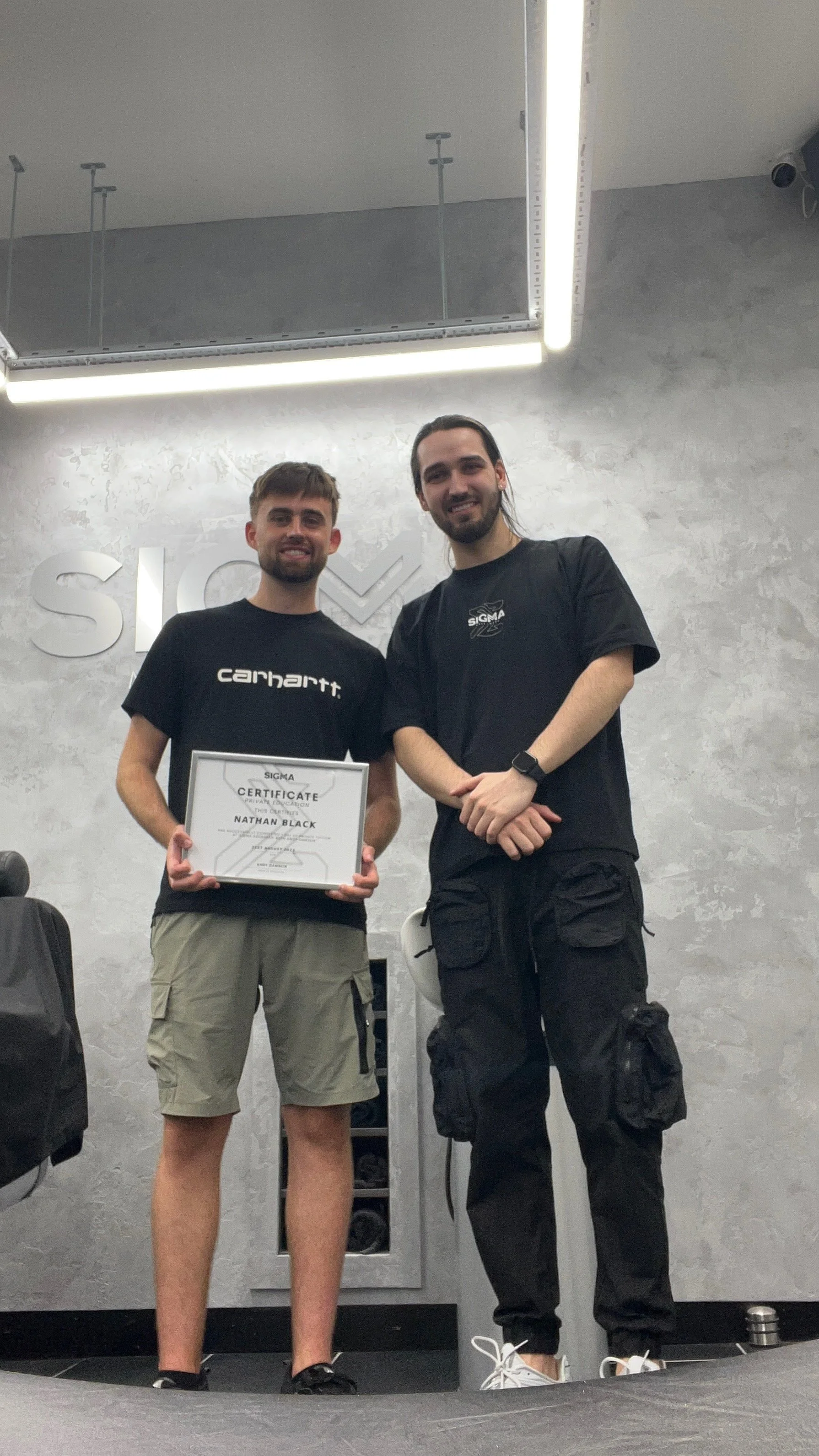 Two young men standing side by side indoors, one holding a framed certificate. They are smiling and wearing black t-shirts, with one in khaki shorts and the other in black cargo pants. The background has a textured gray wall with metallic letters par