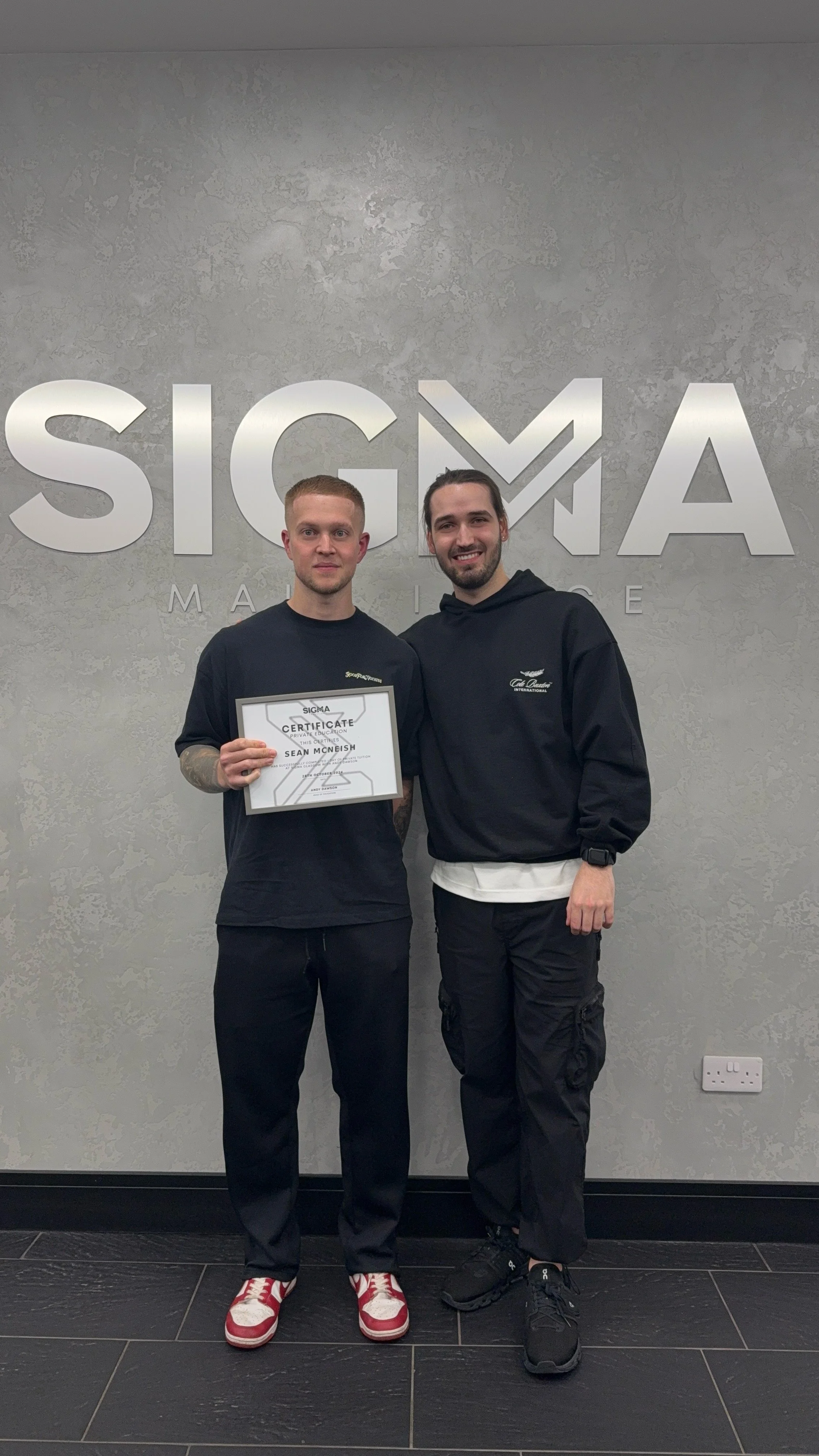 Two young men standing in front of a wall with the Sigma Macing logo, one holding a framed certificate, smiling.
