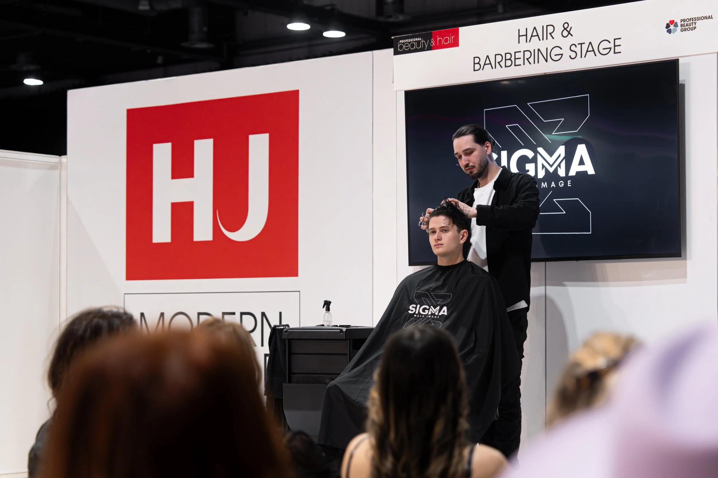 A hairstylist cutting a young man's hair on a stage at a beauty and hair conference, with a large screen and banners displaying HJ Modern and Sigma, in front of an audience.