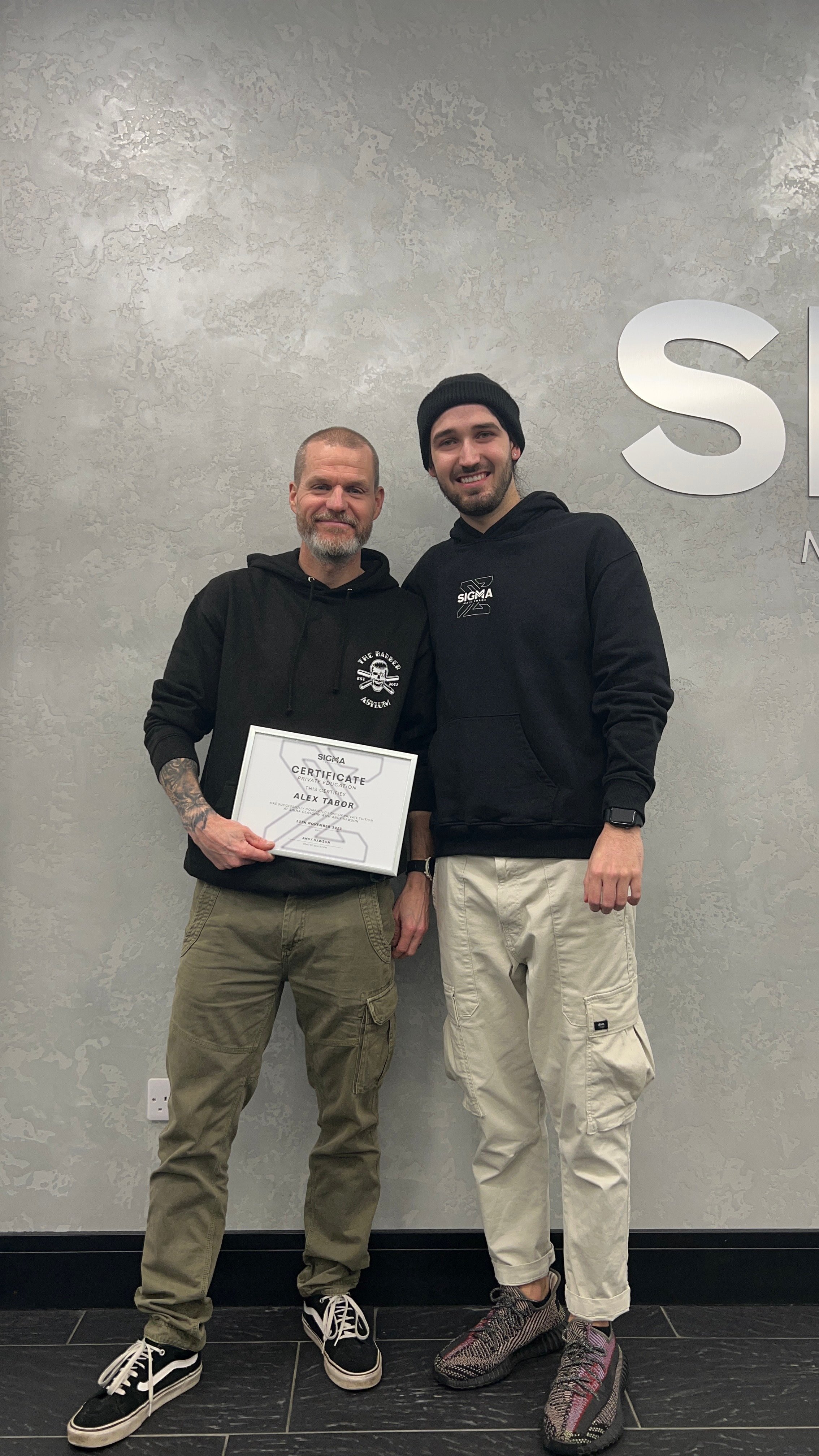 Two men standing against a gray textured wall holding a certificate, smiling. The man on the left has a beard and tattoos, the man on the right wears a beanie and has a beard.