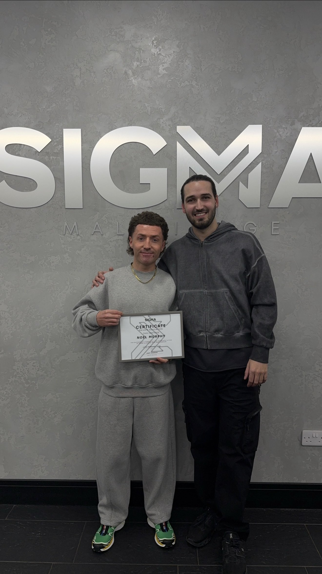 Two men standing in front of a Sigma logo on a gray wall, one holding a certificate. The man on the left is wearing a gray sweatshirt and sweatpants, the man on the right is in a dark hoodie and pants.