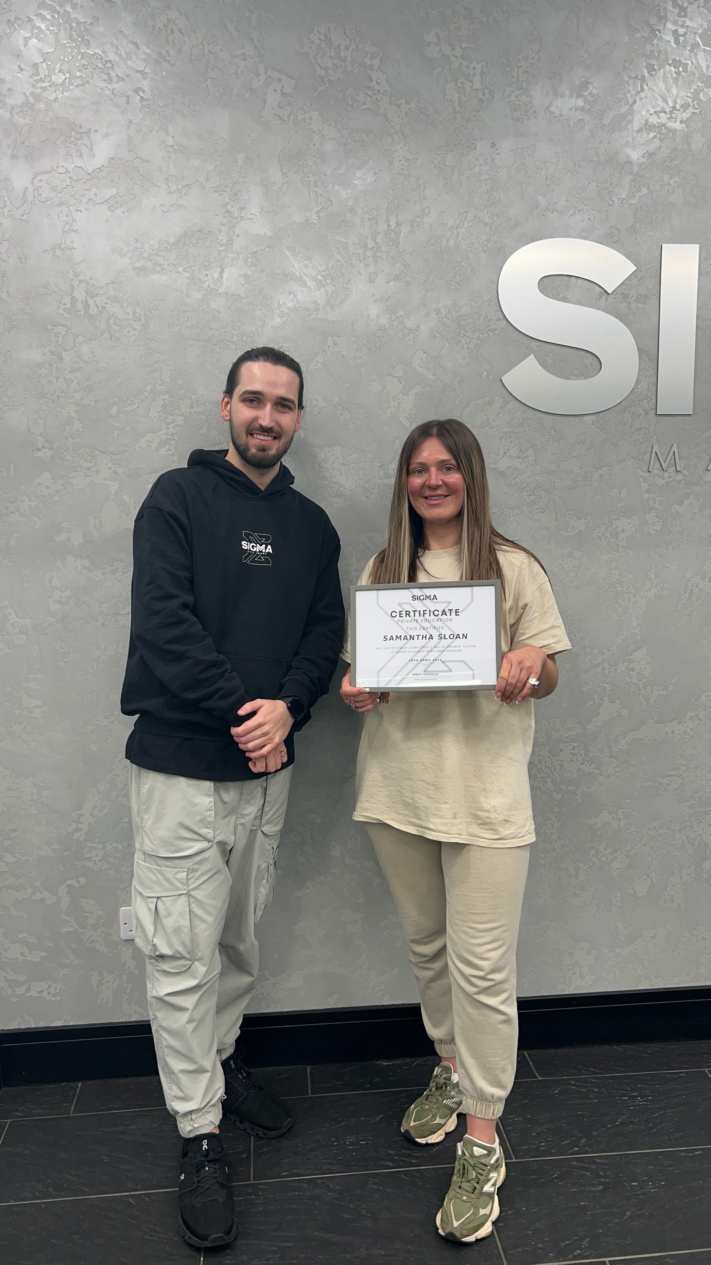 A woman holding a certificate standing next to a man, in front of a gray wall with a partial sign reading 'S I'.