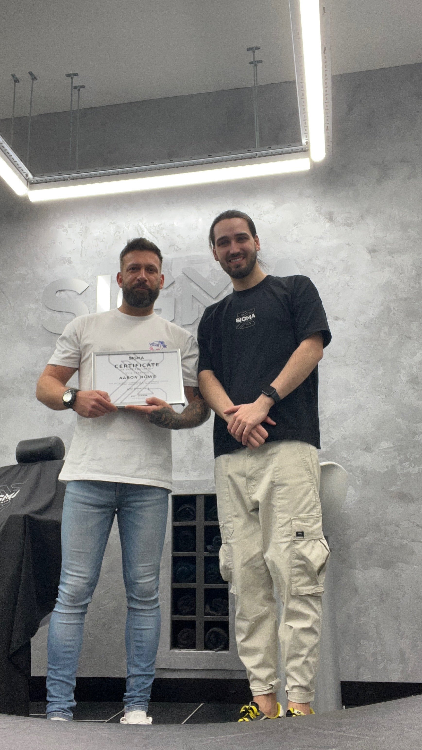 Two men standing indoors, one holding a certificate. They are smiling. The setting appears to be a modern office or showroom with a textured wall background and a large ceiling mirror.