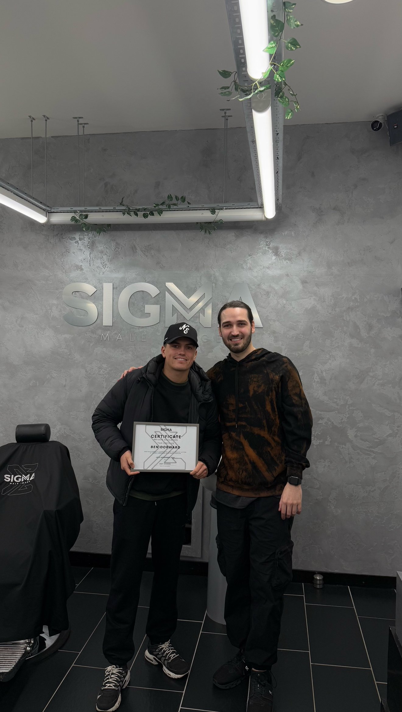 Two young men standing together in a modern office space, smiling at the camera. The man on the left is holding a certificate. The Sigma logo is visible on the wall behind them and on the chair cover.