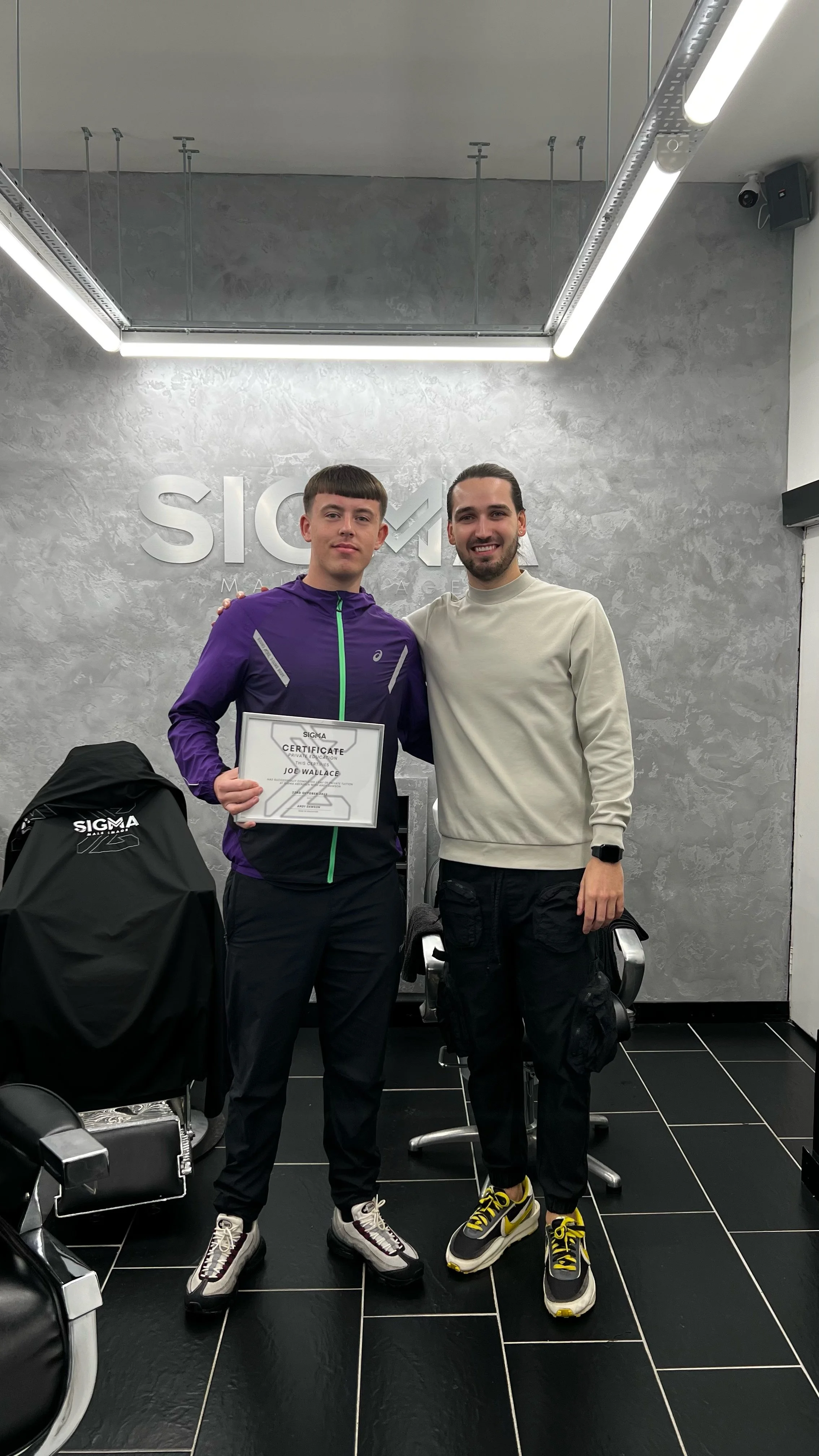 Two young men standing together in a fitness center, one holding a certificate. The background features the SIGMA logo on the wall, and fitness equipment is visible around them.